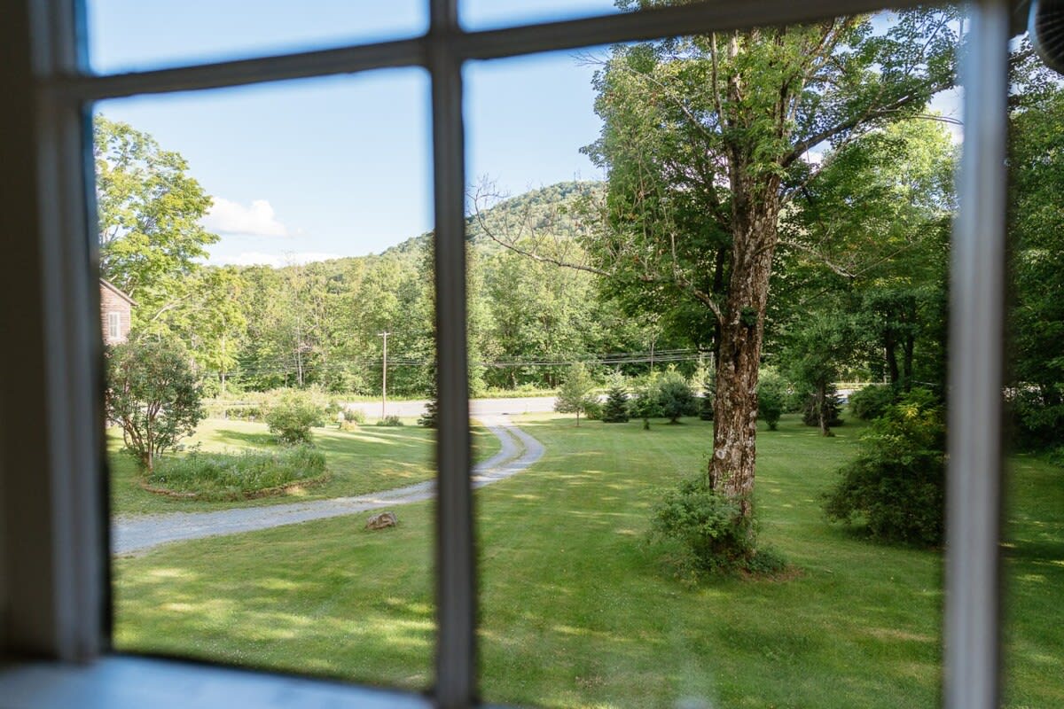 A scenic view from a window showcases a lush green lawn with a winding gravel driveway, surrounded by mature trees and distant hills under a clear sky. The window panes frame the serene rural landscape.