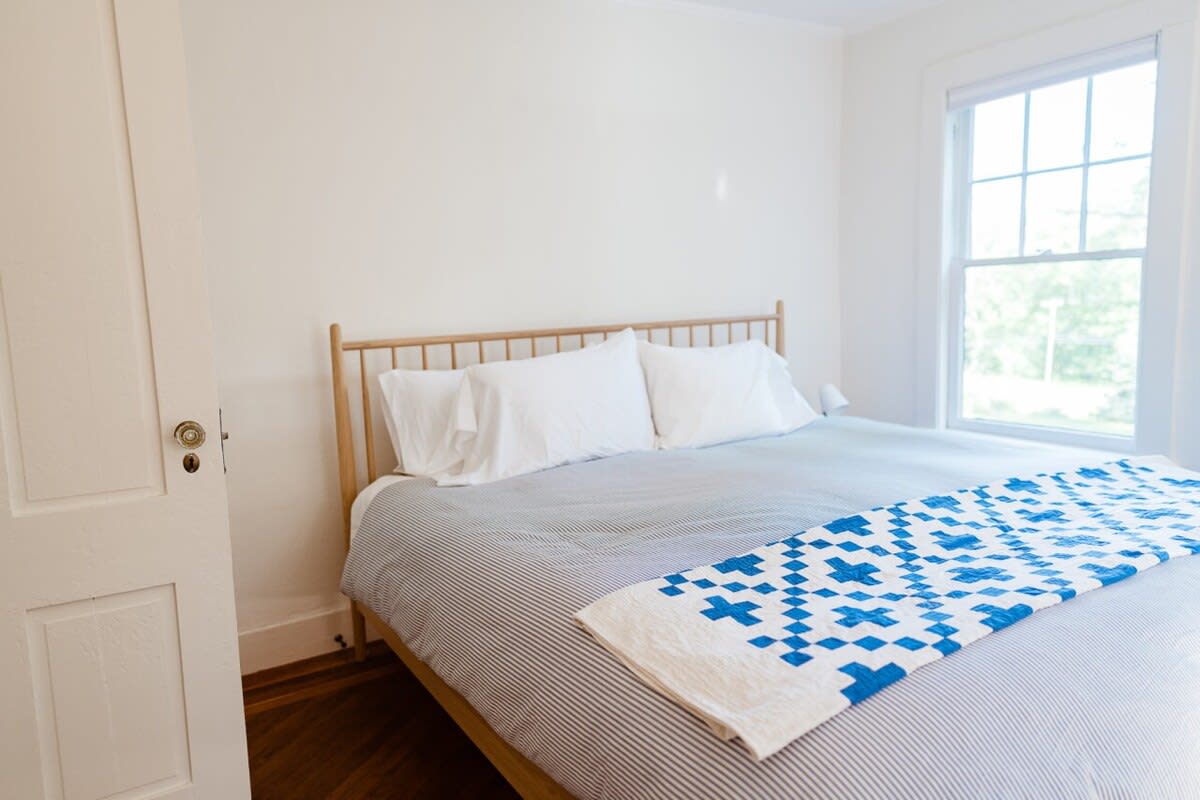 A bright bedroom features a light wood spindle bed frame with white bedding, a striped duvet, and a blue and white patterned quilt. A large window with multiple panes offers natural light, and a white door with a vintage doorknob is visible on the left.