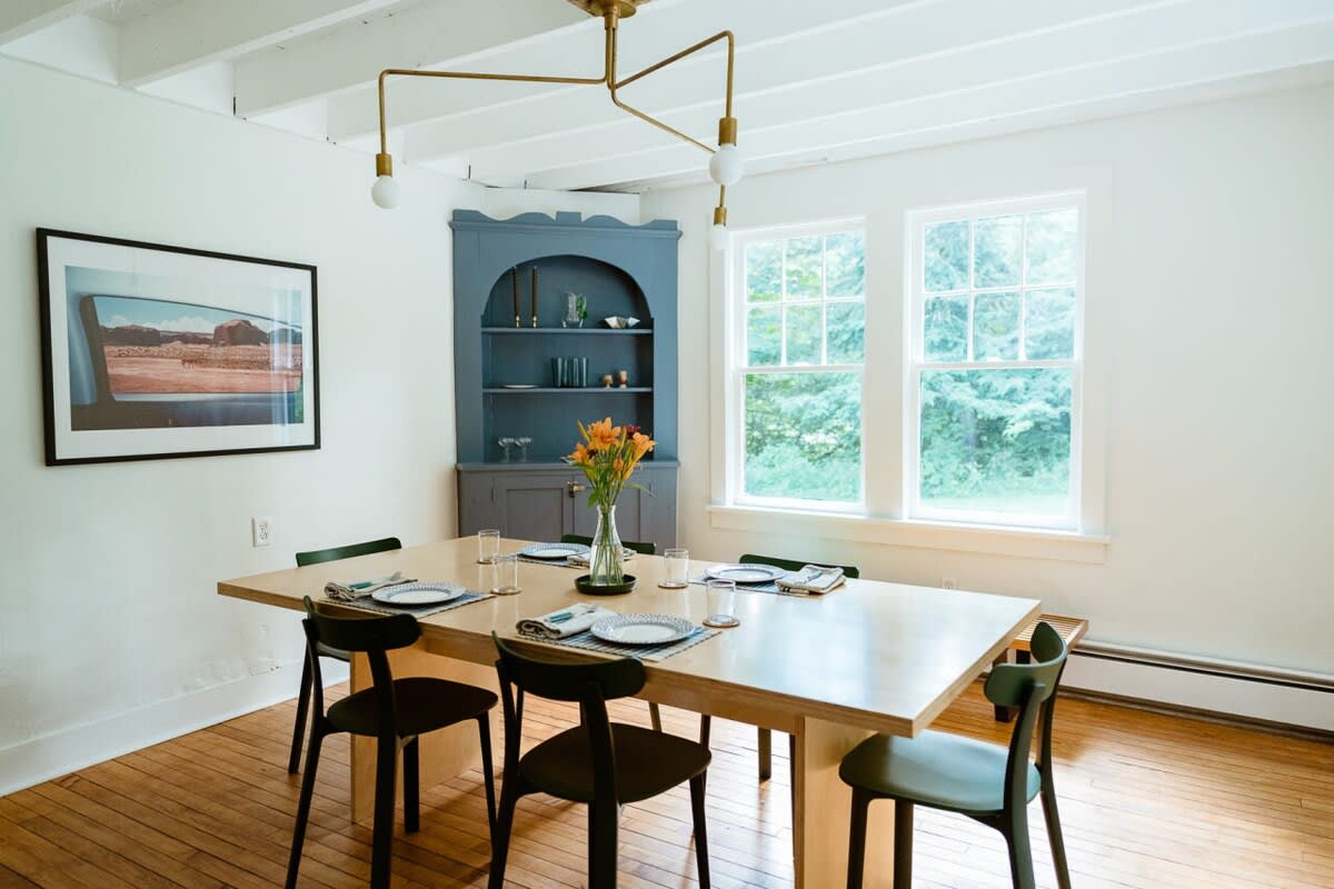 Bright dining room with a light wood table and modern black chairs, featuring a built-in blue corner cabinet and large windows overlooking green foliage.