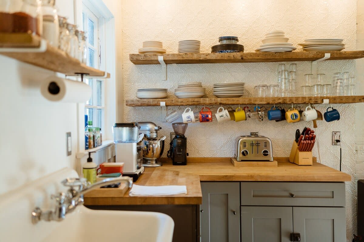 A bright kitchen featuring light wood countertops, open shelving with stacked dishes and hanging mugs, and stainless steel appliances. A white farmhouse sink is visible in the foreground, alongside a toaster and coffee grinder.