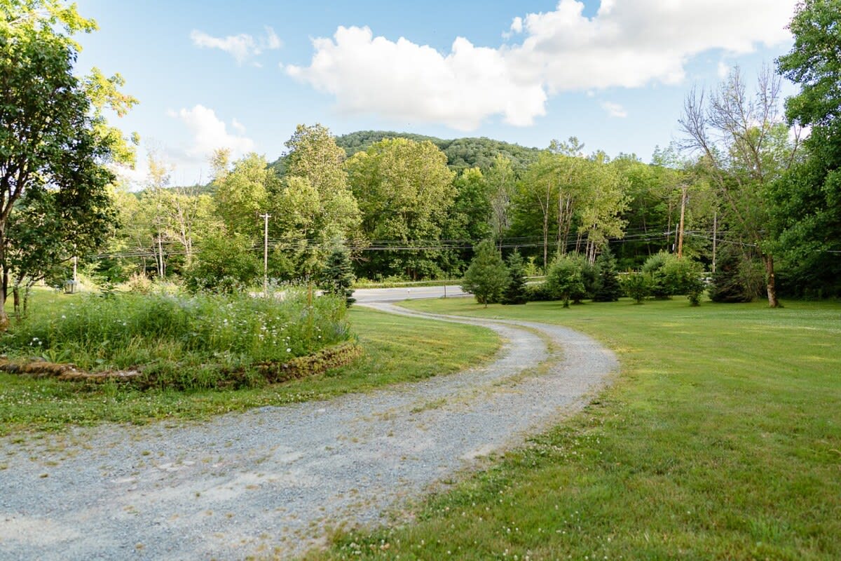 A winding gravel driveway cutting through a well-maintained green lawn, bordered by wildflowers and mature trees, with a distant view of a forested hill under a blue sky.