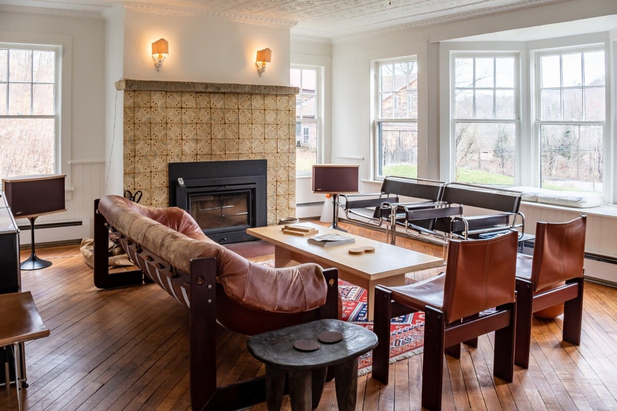 Bright living room with hardwood floors, a large brown leather sofa, and a fireplace with a tiled surround, featuring bay windows overlooking a wooded landscape.
