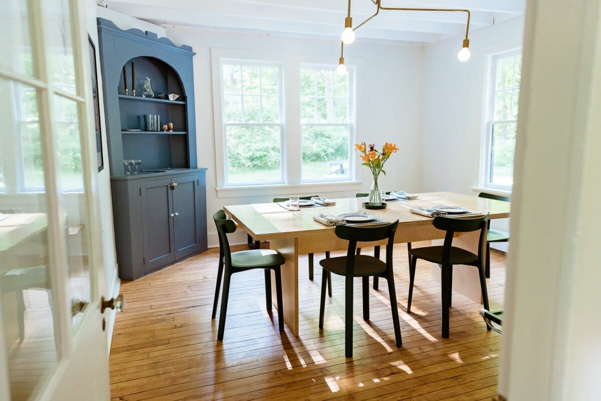 A bright dining room featuring a large light wood dining table with six dark green chairs, set against a backdrop of multiple windows. A dark blue built-in hutch with arched shelving adds a pop of color.