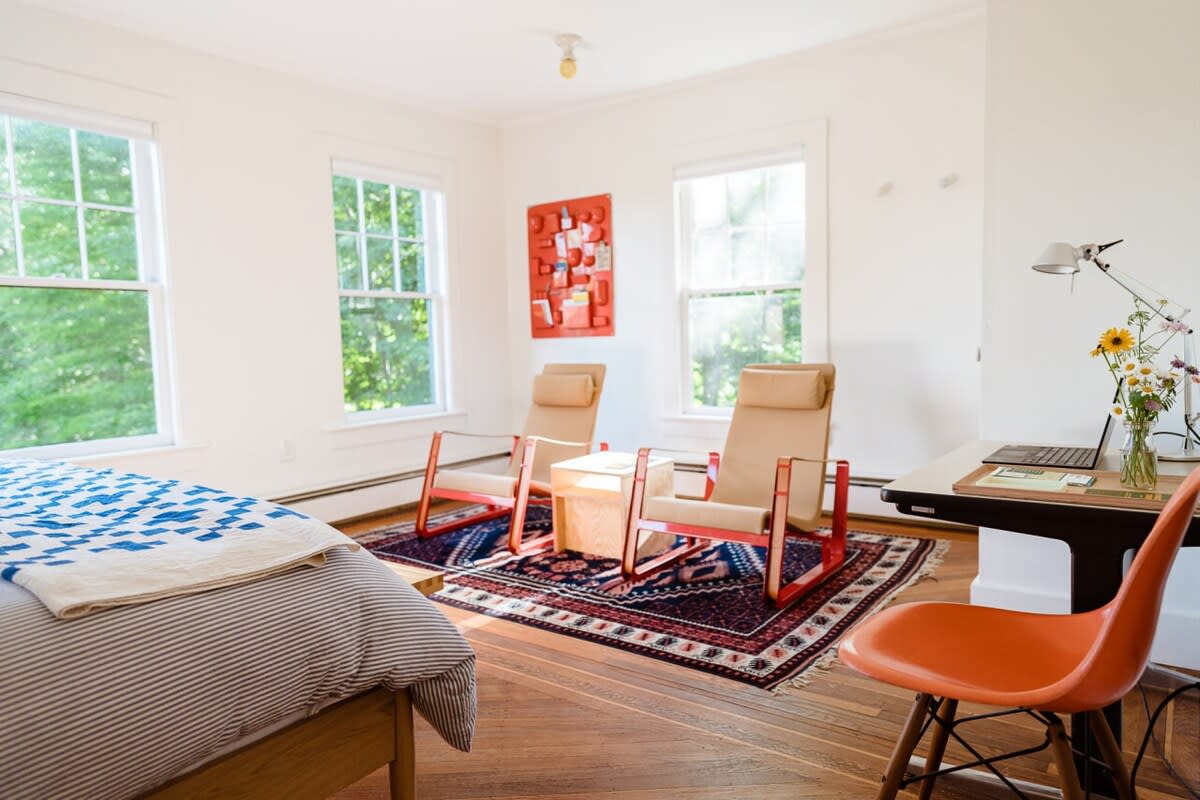 A spacious bedroom features a bed with a blue and white quilt in the foreground, leading to a seating area with two modern tan armchairs with red frames, a small wooden table, and a desk with an orange chair. Large windows provide ample natural light, and a vibrant red abstract artwork hangs on the wall.