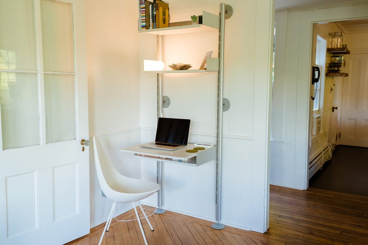 A modern wall-mounted desk and shelving unit with a laptop, positioned in a bright corner next to a white paneled door. A white contemporary chair is pulled up to the desk, offering a dedicated workspace.