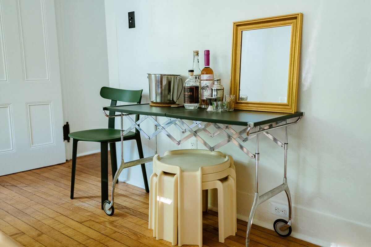 A mobile bar cart with a dark green top, holding various bottles and a metal ice bucket, is positioned against a white wall. A green chair and stackable stools are nearby, creating a functional beverage station.