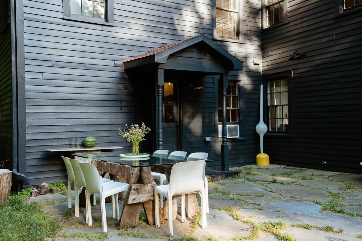 Stone patio area next to the dark gray house, featuring a rustic outdoor dining table with a glass top and white chairs, shaded by the house's overhang.