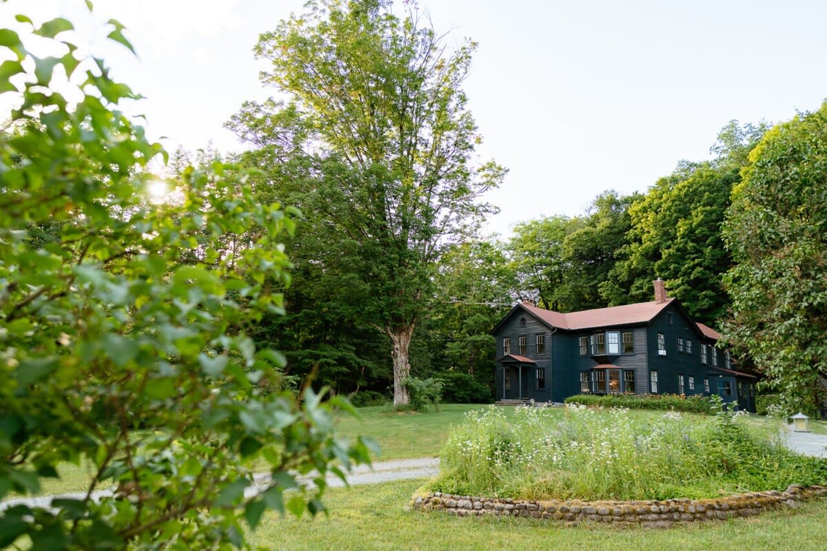 A dark gray farmhouse with a reddish-brown roof nestled among tall green trees, viewed from a grassy lawn with wildflowers and a gravel driveway.