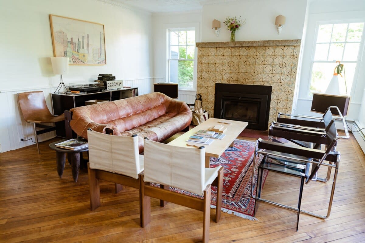 A spacious living room with a distressed leather sofa, a light wood coffee table, and various seating options arranged around a tiled fireplace. Hardwood floors are partially covered by a patterned area rug.