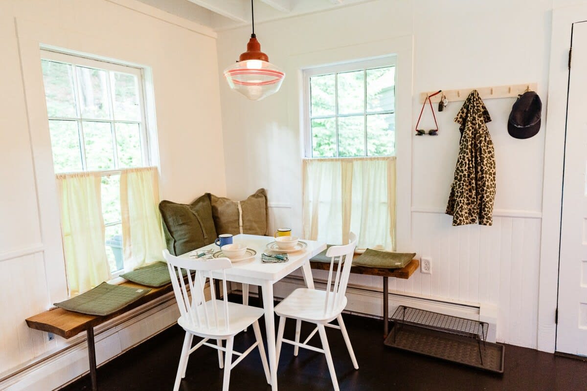 A cozy breakfast nook with a white square dining table and two white spindle-back chairs, flanked by built-in wooden benches with olive green cushions. Large windows with sheer yellow curtains overlook the outdoors.