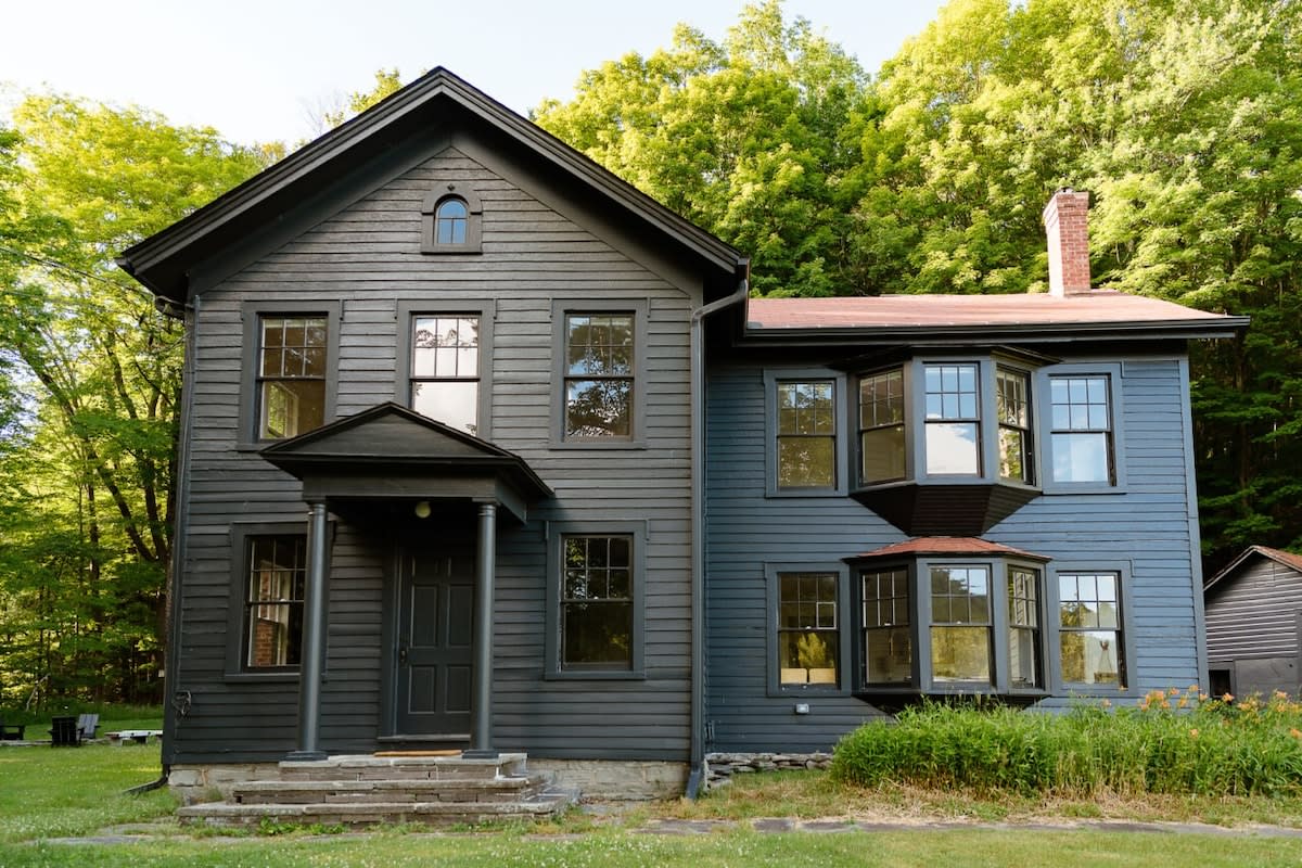A dark gray, two-story farmhouse with a gabled roof and multiple windows, featuring a covered front porch with columns, set against a backdrop of lush green trees.