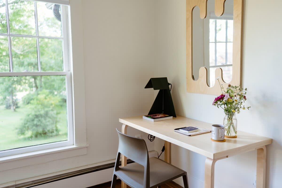 A simple and bright workspace featuring a light wood desk, a modern gray chair, and a black task lamp. A large window offers a view of lush greenery outside, providing natural light.