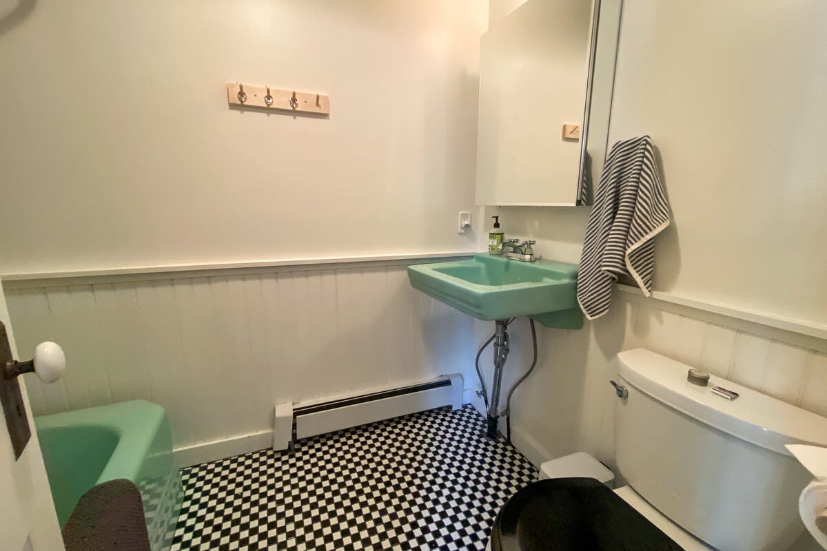 A vintage-style bathroom features a mint green pedestal sink and a matching bathtub, set against white wainscoting and black and white checkered floor tiles. A toilet is visible on the right, and a mirror hangs above the sink.
