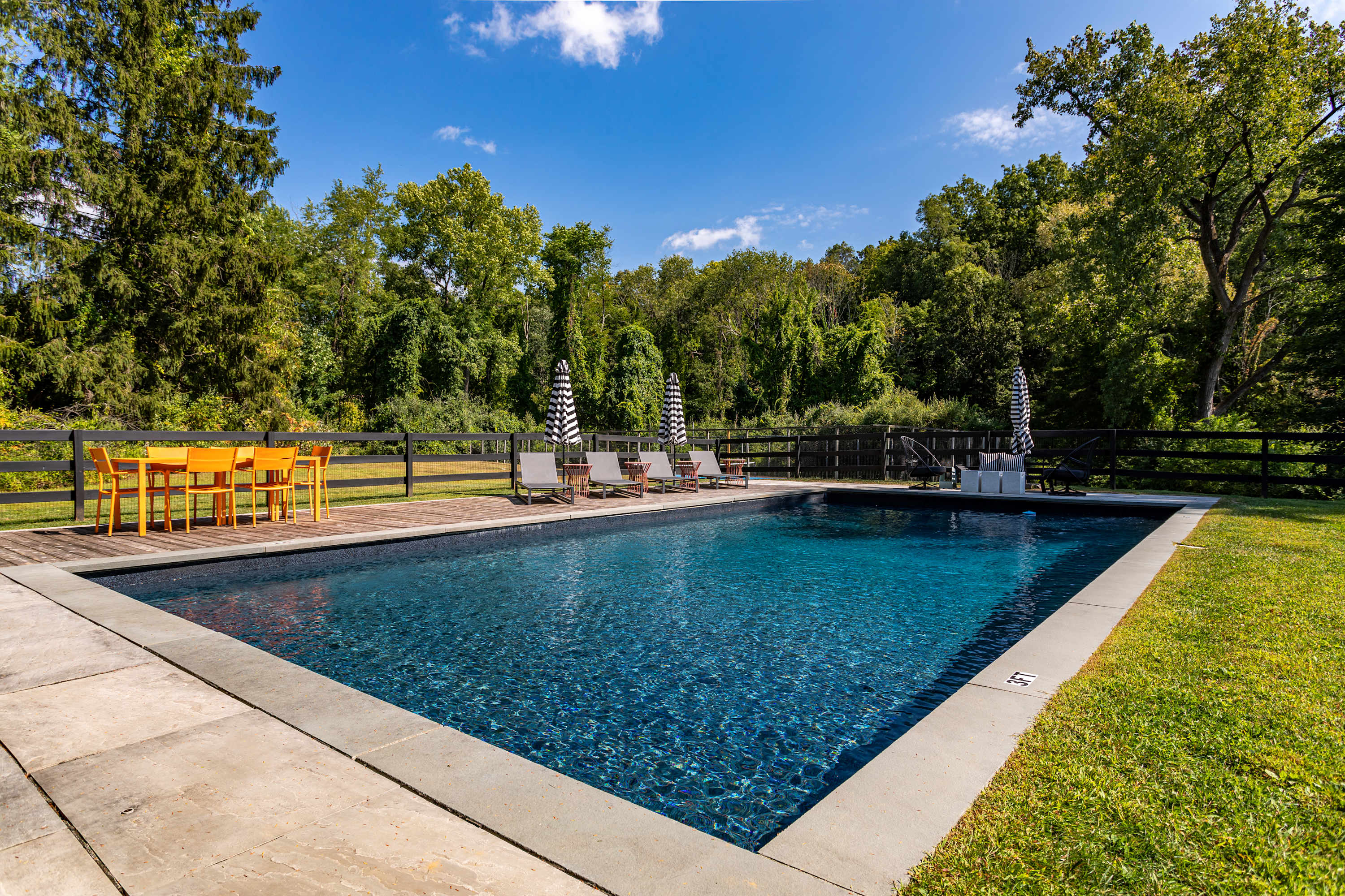 Expansive view of a dark blue swimming pool with a stone coping, flanked by a wooden deck with an orange dining set and lounge chairs with striped umbrellas, all set against a backdrop of dense green trees.