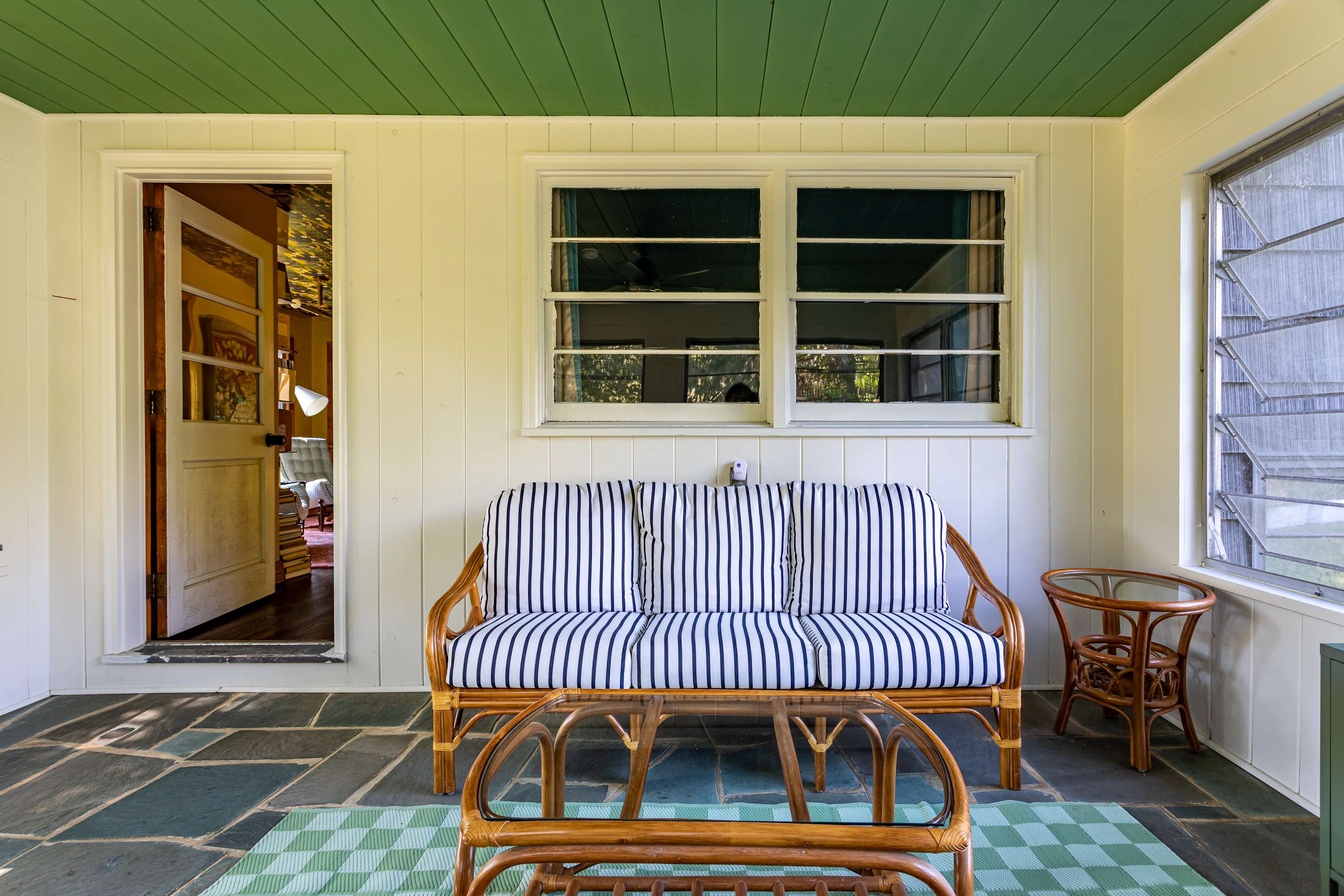 A cozy screened porch with a green slatted ceiling, white paneled walls, and a dark slate floor. It features a rattan sofa with striped cushions, a glass-top coffee table, and a view into an interior room through an open door.