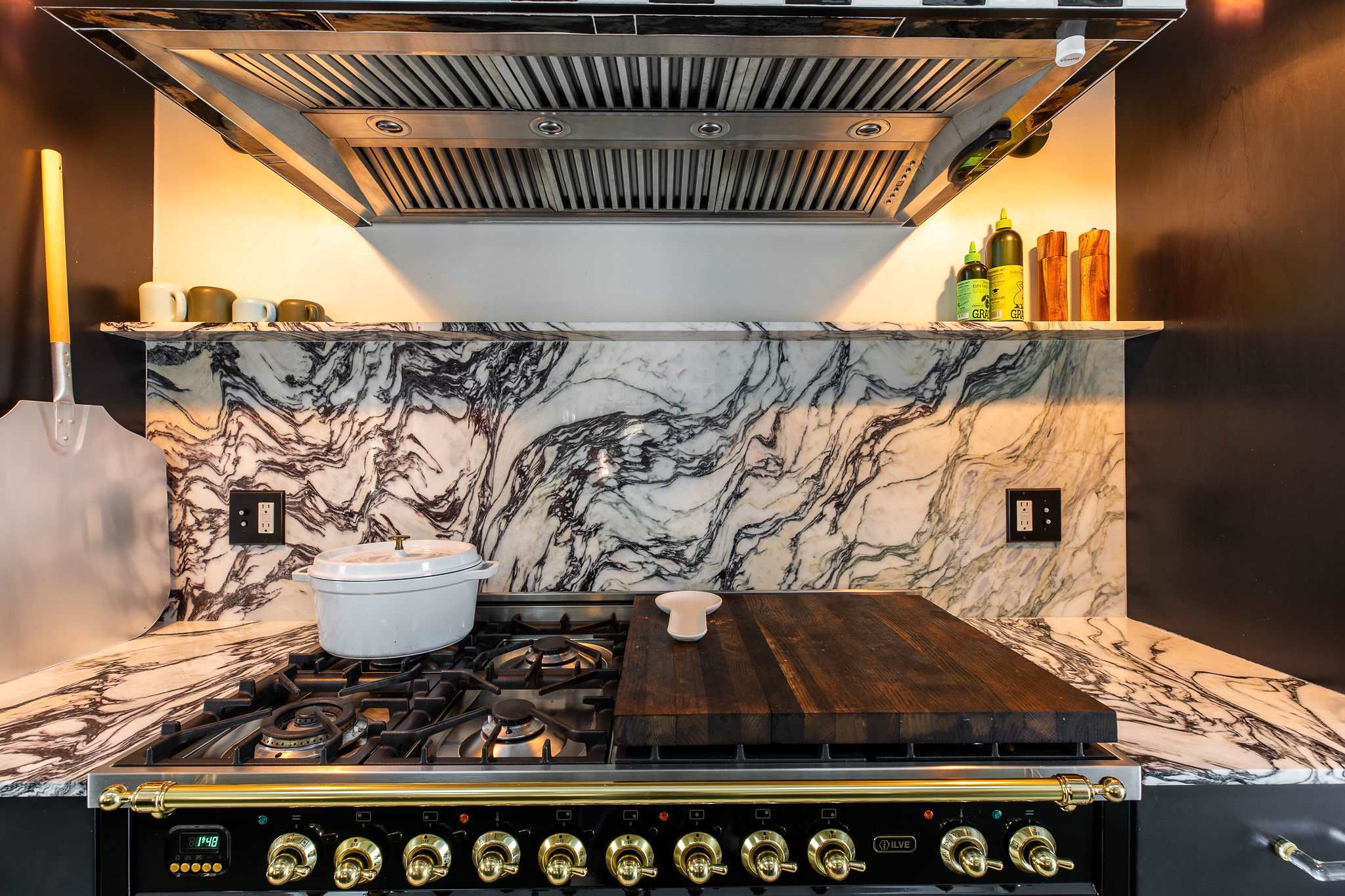 A detailed view of a professional-style black and gold range with a large wooden cutting board, set against a dramatic white marble backsplash with dark veining, under a stainless steel range hood.