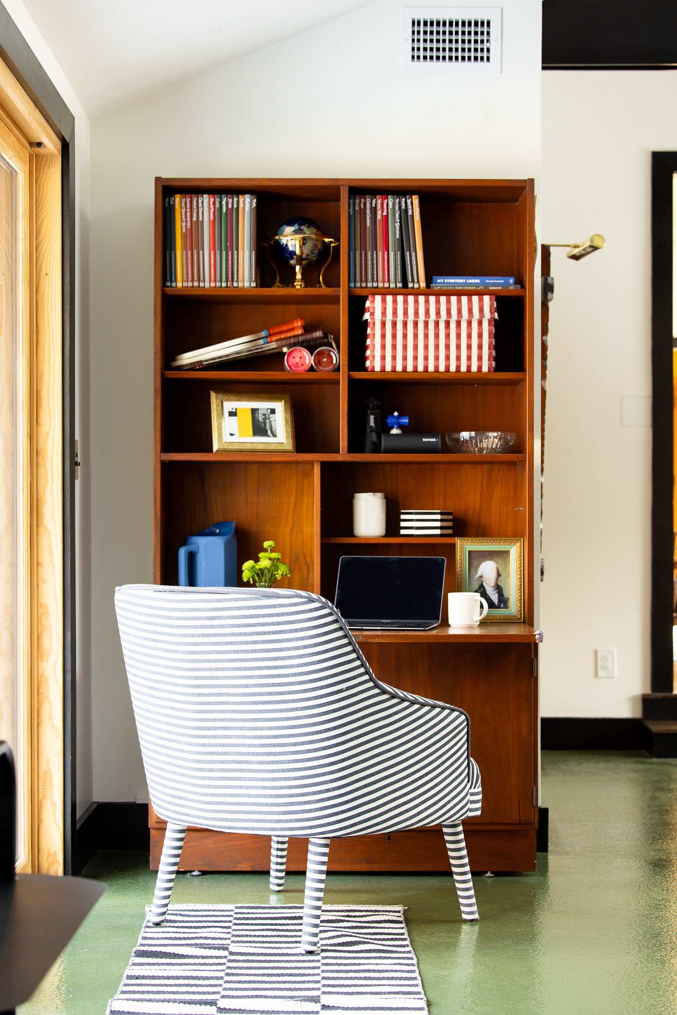Dedicated workspace featuring a wooden bookshelf filled with books and decor, a striped upholstered chair, and a laptop on a pull-out desk. The floor is a glossy green, and natural light comes from a nearby door.
