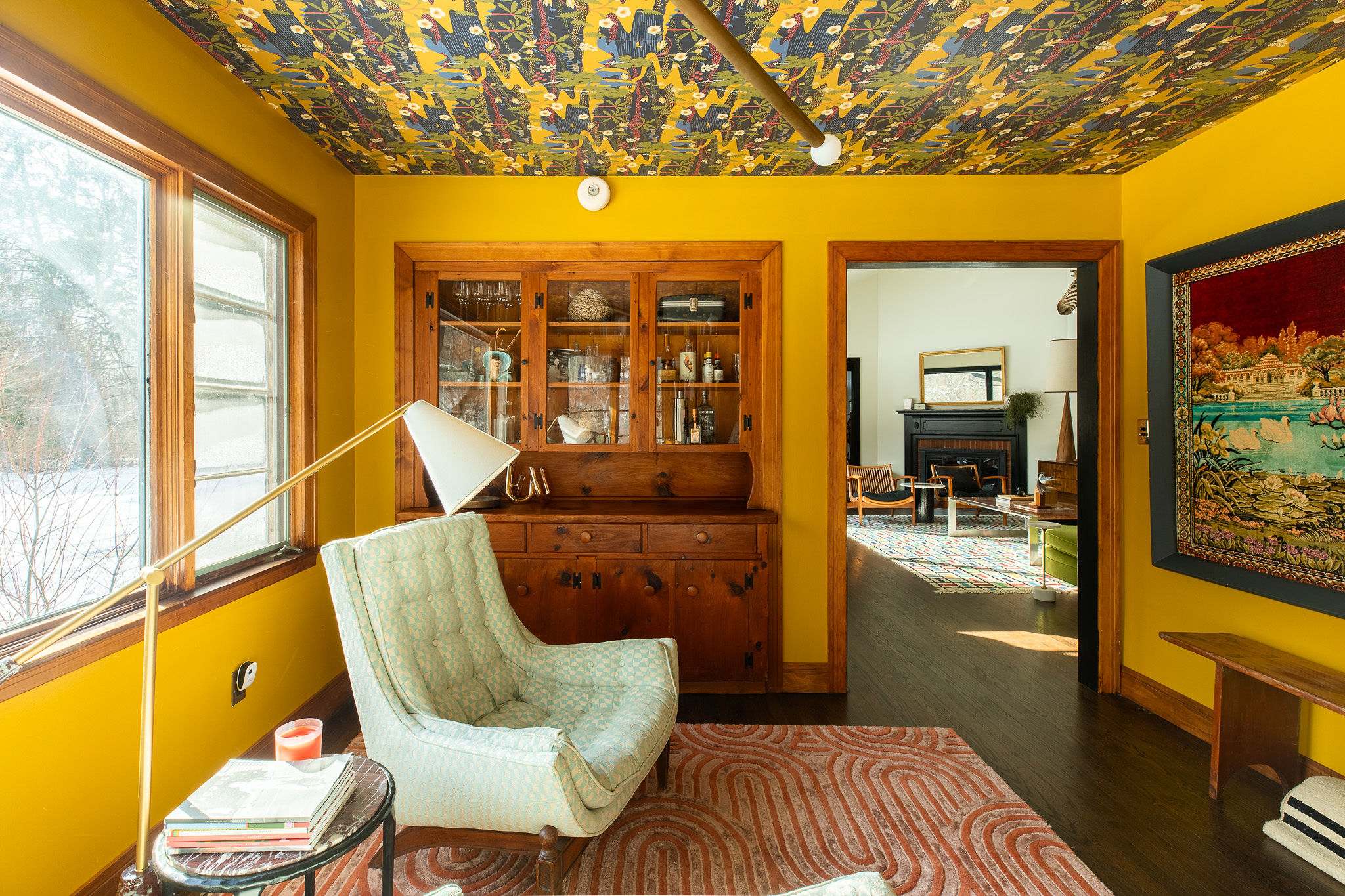 A cozy room with bright yellow walls and a patterned ceiling features a vintage armchair, a wooden hutch filled with items, and a floor lamp, with a view into another room.
