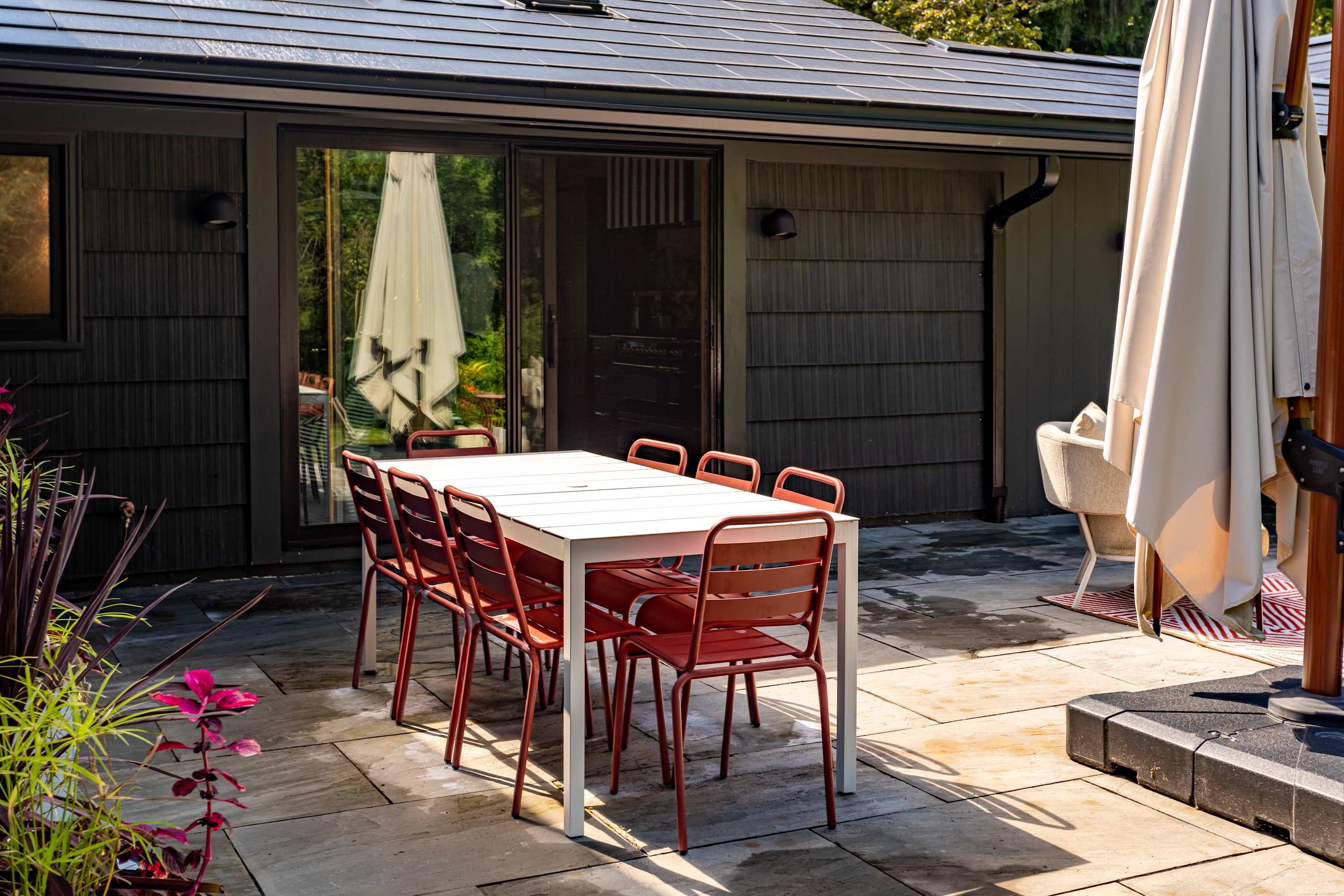 An outdoor dining area on a stone patio next to a dark-sided building with sliding glass doors, featuring a white rectangular dining table with red chairs and a large beige patio umbrella.