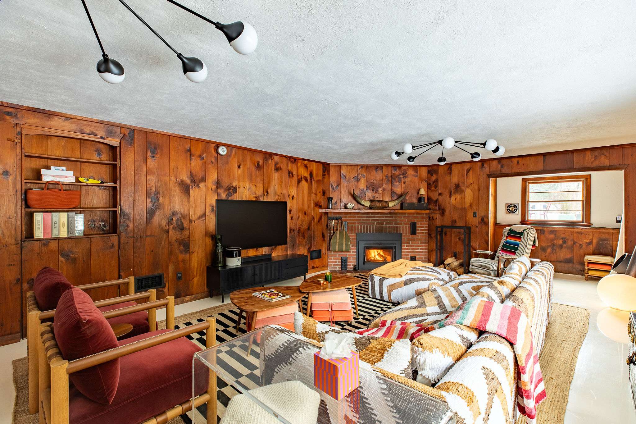 Cozy living room featuring warm wood-paneled walls, a brick fireplace with a wood-burning insert, and a large sectional sofa adorned with a colorful patterned throw blanket. A modern black chandelier hangs from the ceiling.