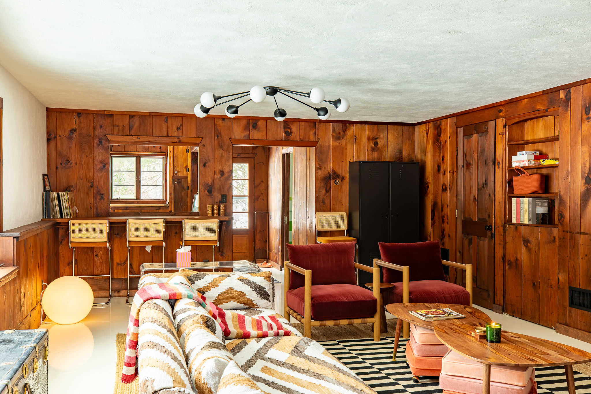 Expansive living room with rustic wood-paneled walls, a large patterned sectional sofa, and two red velvet armchairs, centered around a black and white striped rug. A bar area with three cane-back stools is visible in the background.