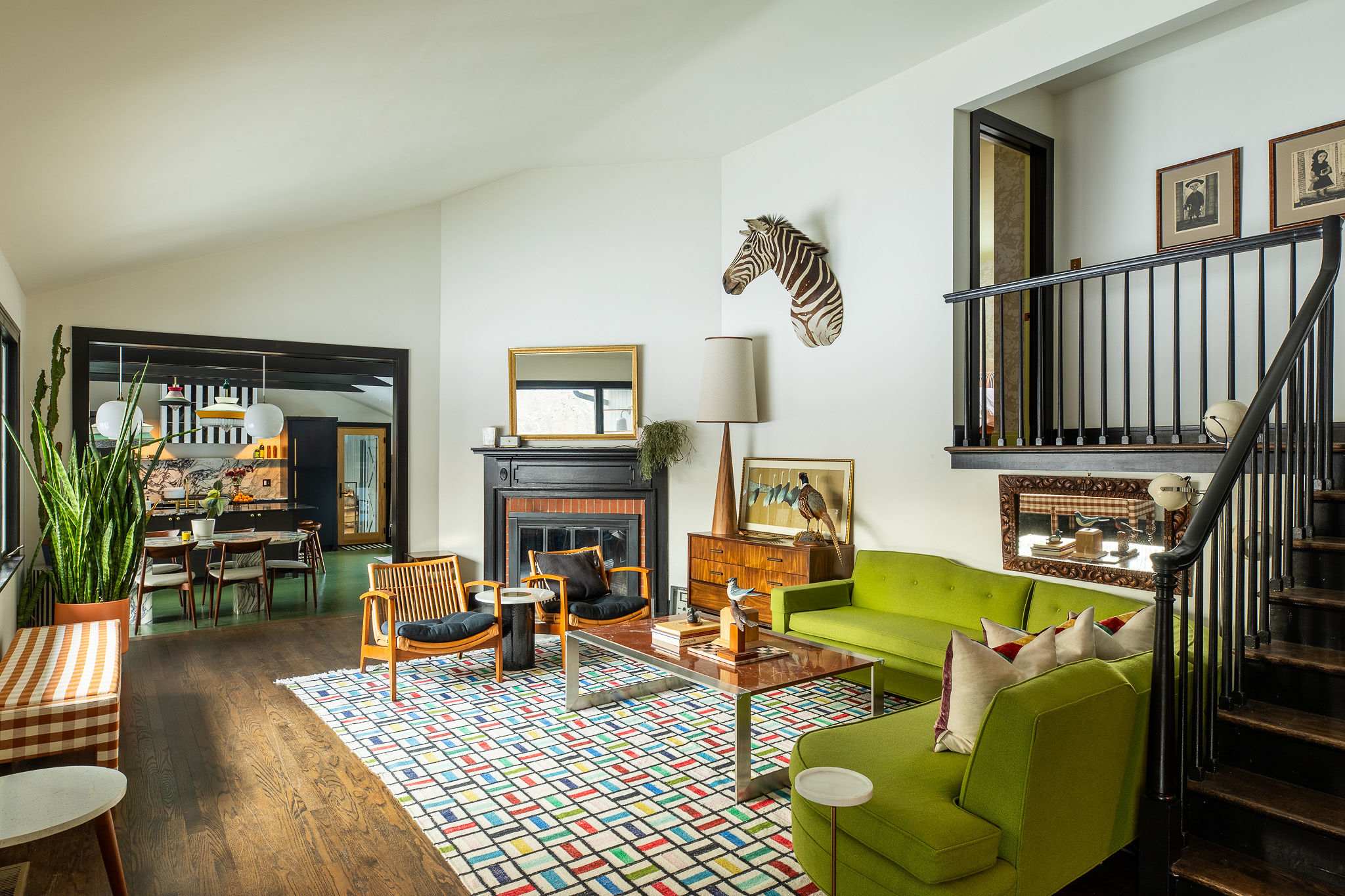 A vibrant living room features a lime green sectional sofa, two rattan armchairs, a colorful geometric rug, and a fireplace with a black mantel, all under a high ceiling with a zebra head wall decor.