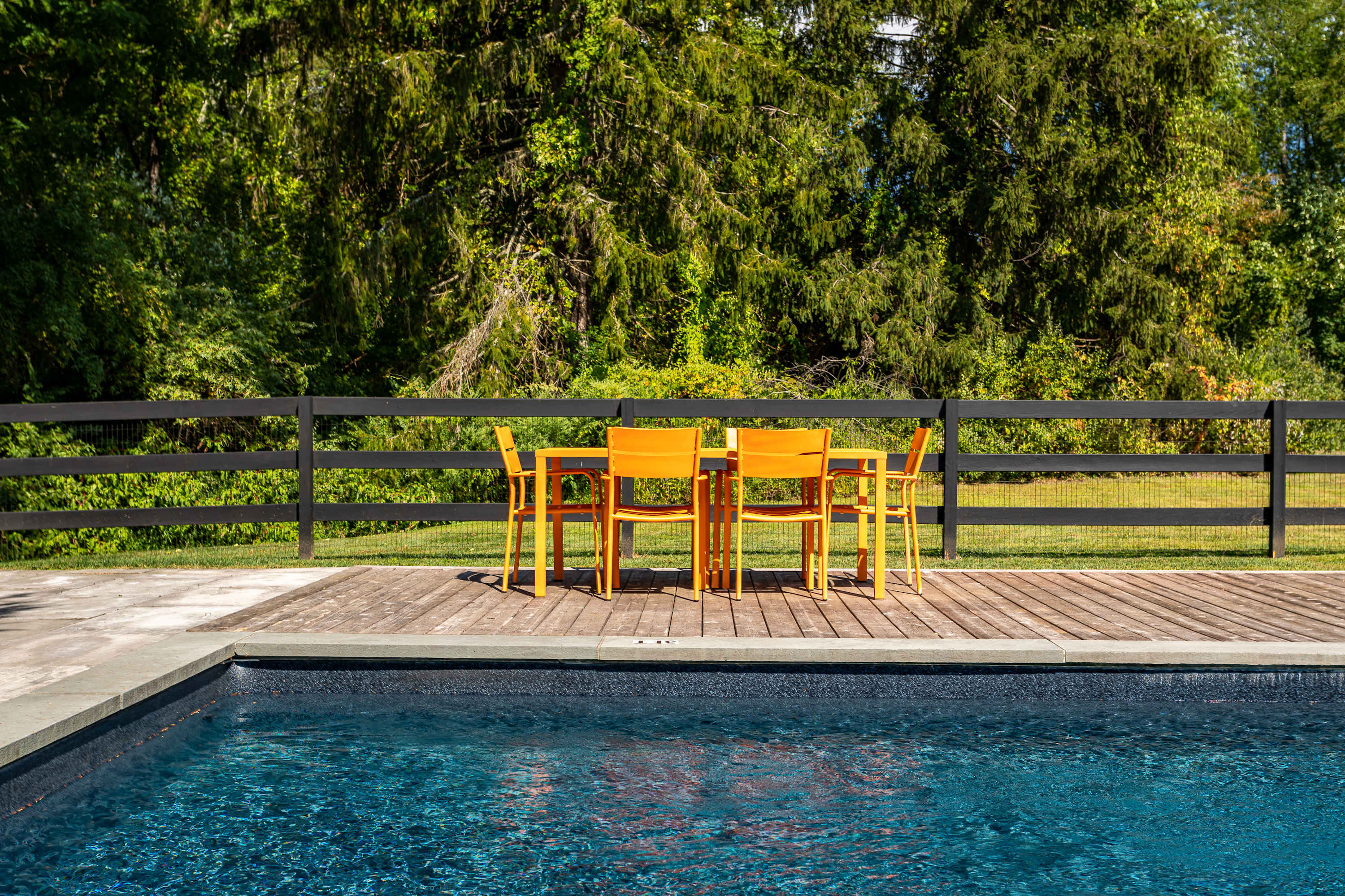 A vibrant orange outdoor dining set with six chairs sits on a wooden deck next to a dark blue swimming pool, surrounded by a black fence and lush green trees under a bright sky.