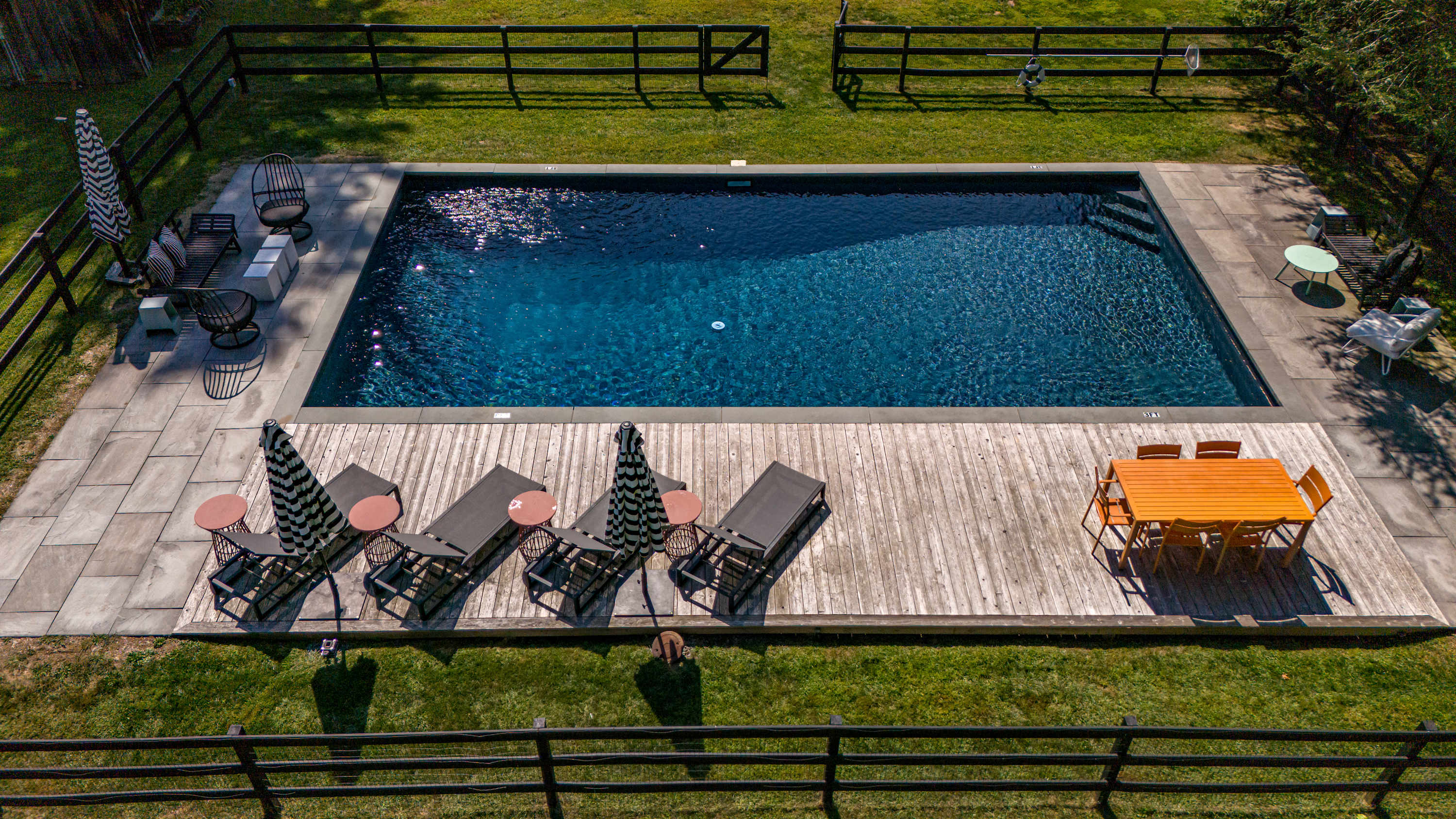 An aerial shot of a rectangular swimming pool with dark blue water, surrounded by a light-colored stone deck and a wooden deck area. The deck is furnished with lounge chairs, umbrellas, and an outdoor dining set, all enclosed by a black fence.