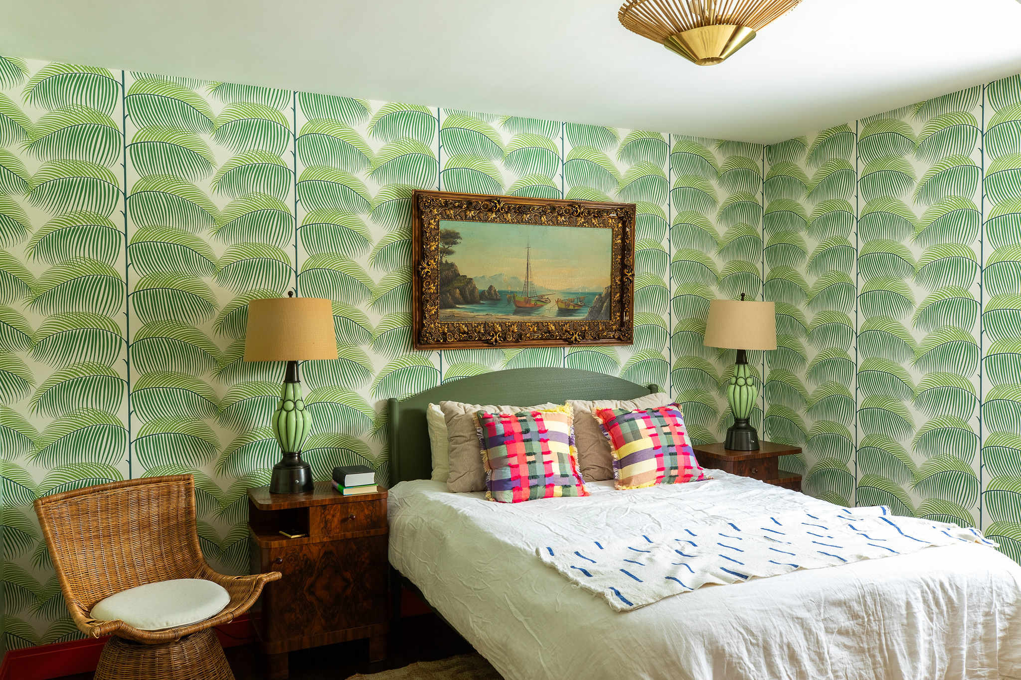 Vibrant bedroom adorned with green palm leaf patterned wallpaper, featuring a bed with white bedding and colorful plaid pillows, two wooden nightstands with lamps, and a wicker chair. A framed painting hangs above the bed.