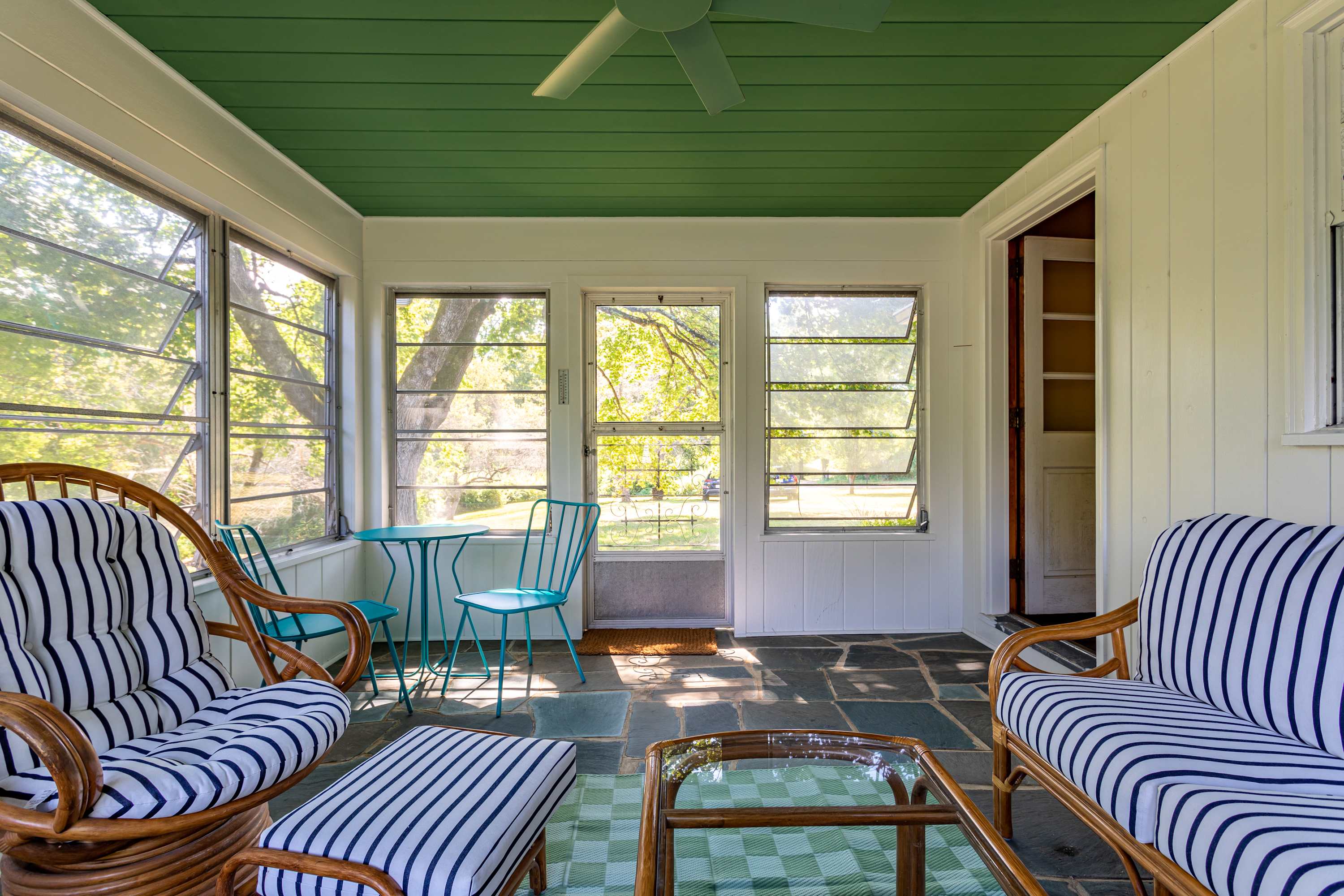 A bright screened porch with a green slatted ceiling, white paneled walls, and a dark slate floor. It features rattan furniture with striped cushions, a small blue bistro set, and large windows overlooking a leafy outdoor scene.