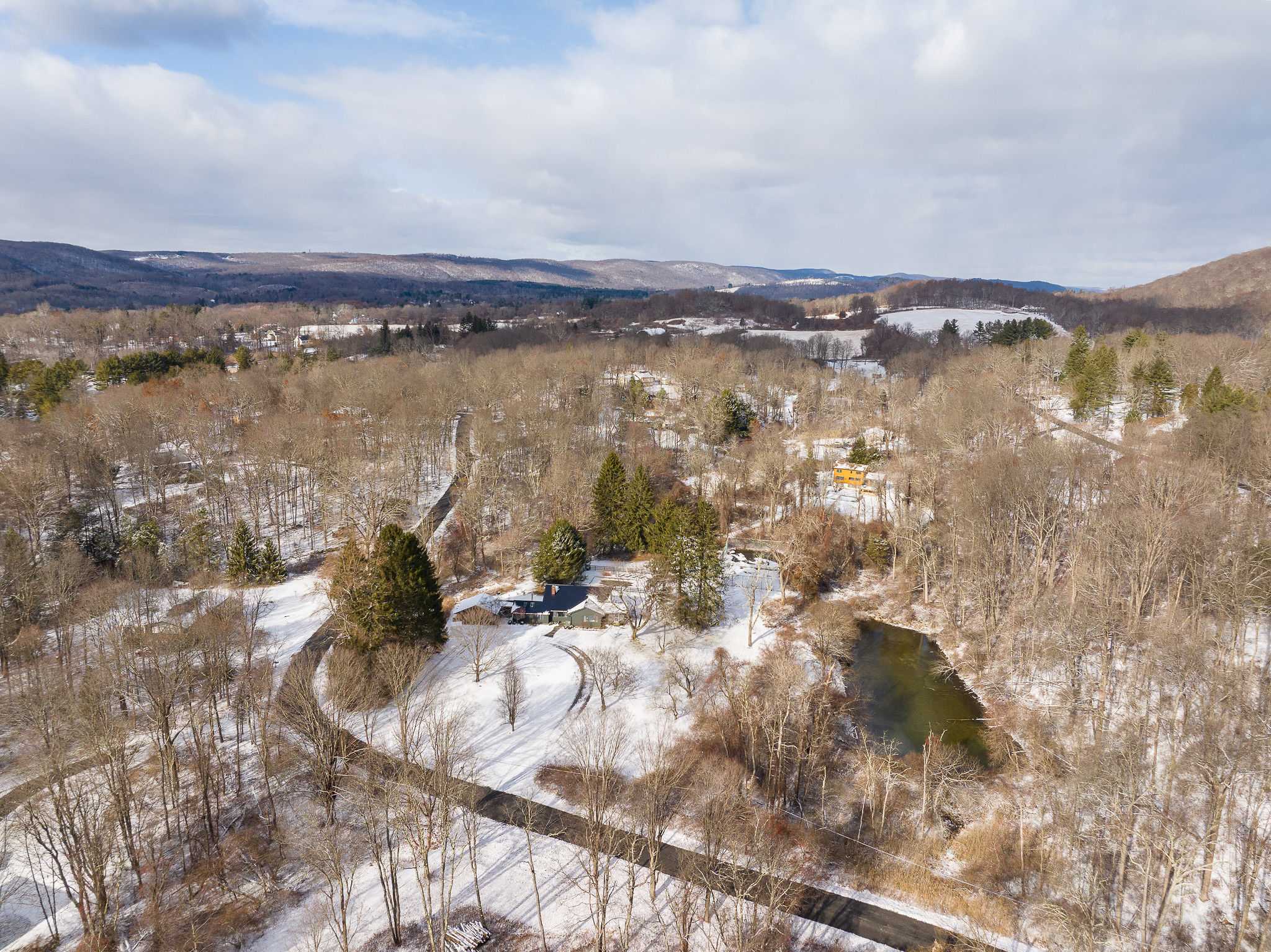 An aerial view of a snow-dusted property featuring a dark-colored house nestled among bare trees, with a winding driveway and a dark pond, all set against a backdrop of rolling hills and a cloudy sky.