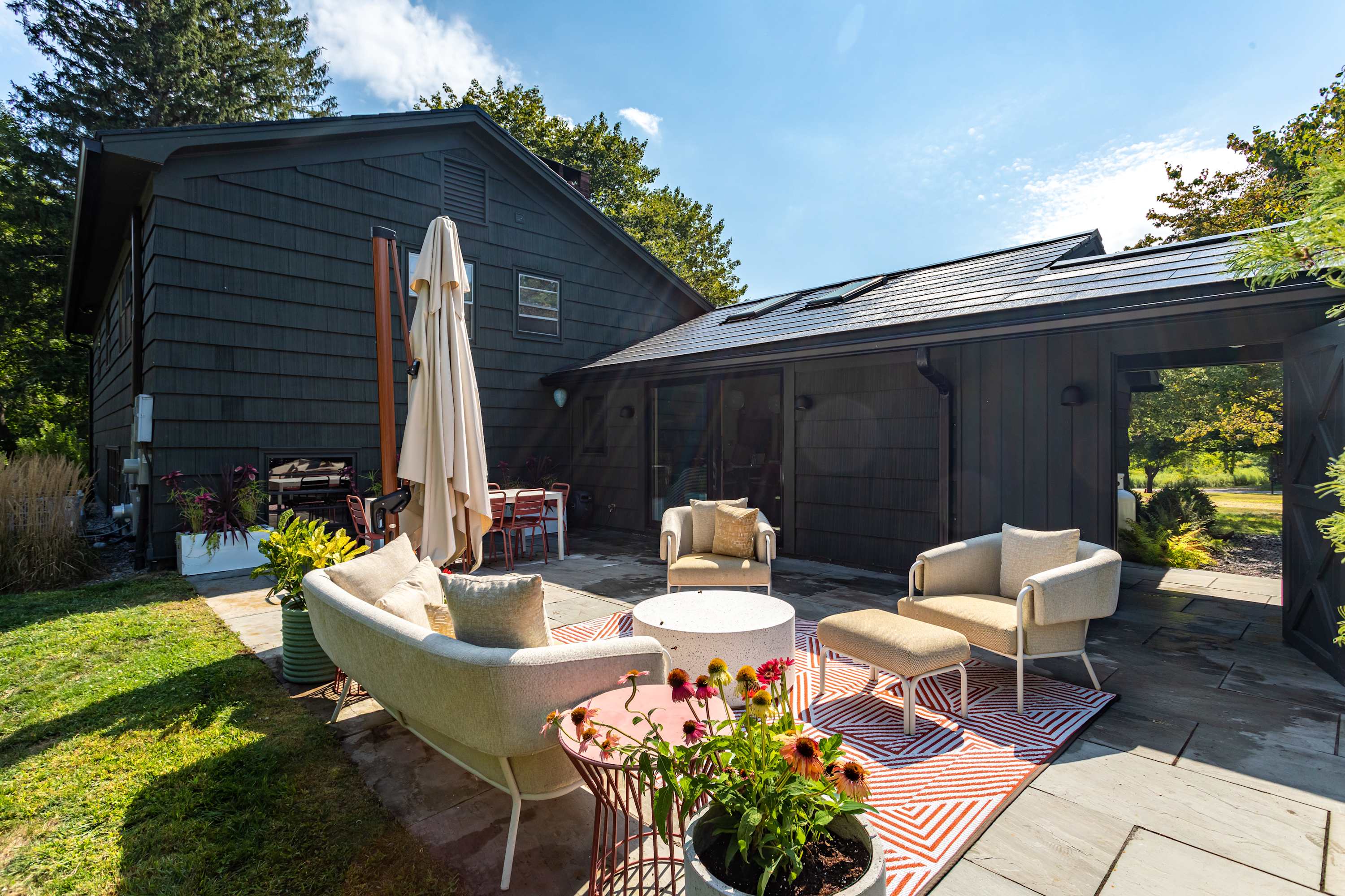 A stylish outdoor patio area with modern seating, including a sofa, armchairs, and a coffee table, is set on a stone surface next to a dark-sided house, with an umbrella providing shade and potted plants adding greenery.