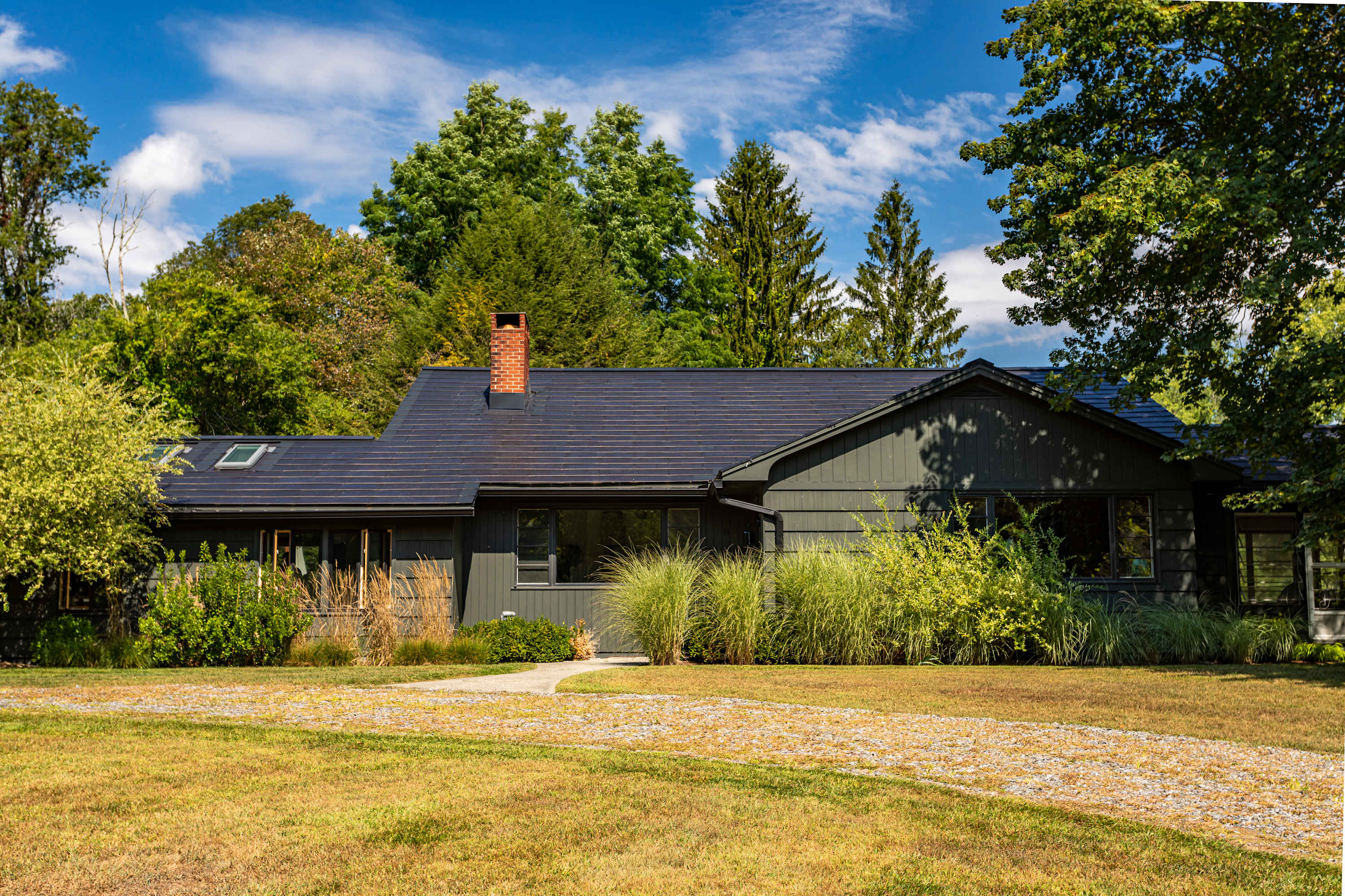 The front exterior of a modern dark-colored house with a dark roof and solar panels, surrounded by lush green landscaping and mature trees. A curved gravel path leads to the entrance, with a well-kept lawn in front.