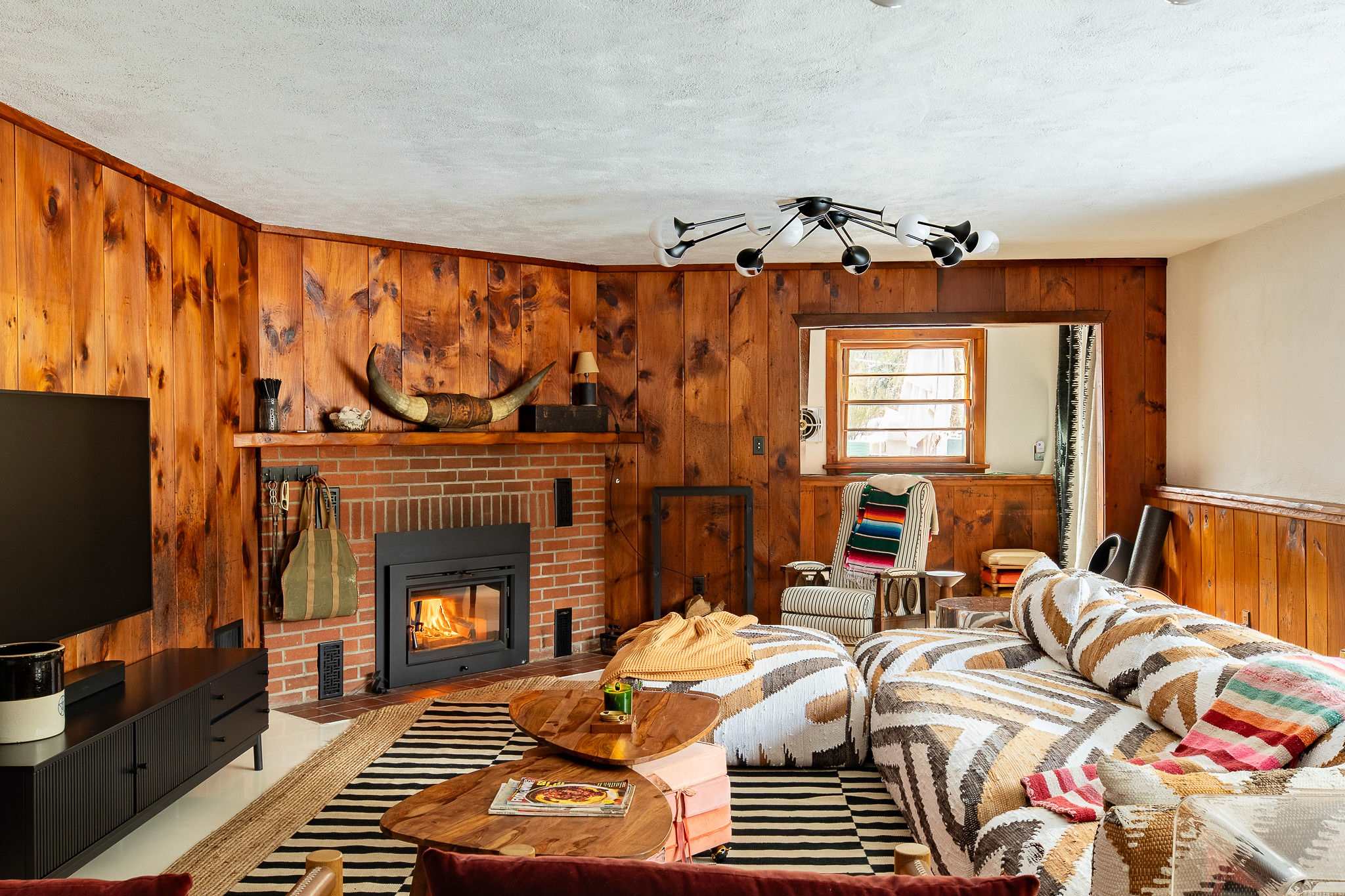A rustic living room features wood-paneled walls and a brick fireplace with a burning fire, topped by a mantel displaying decorative horns. A large, patterned sectional sofa sits on a black and white striped rug, with unique wooden coffee tables in front.
