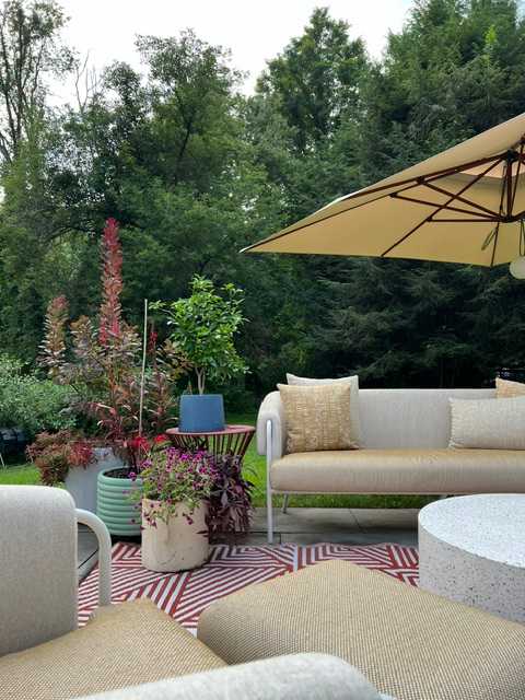 A close-up view of the outdoor patio, showcasing a beige outdoor sofa with decorative pillows, potted plants, and a red and white geometric rug. A large patio umbrella provides shade over the comfortable seating area.