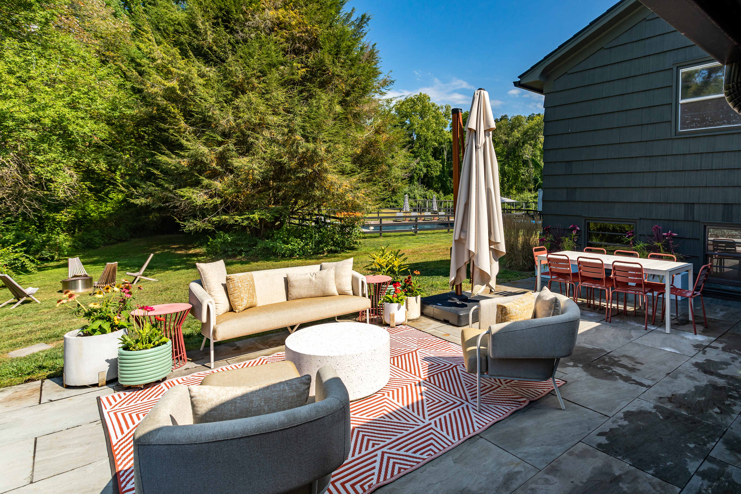 An expansive outdoor patio with a lounge area featuring a beige sofa and armchairs on a geometric rug, and a separate dining area with a white table and red chairs, all under a large umbrella, overlooking a grassy yard and distant pool.