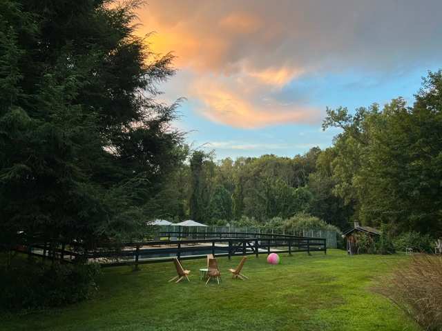 A spacious green yard with a fenced swimming pool area in the background, surrounded by mature trees under a dramatic sunset sky. Several Adirondack chairs and a pink ball are scattered on the lawn.