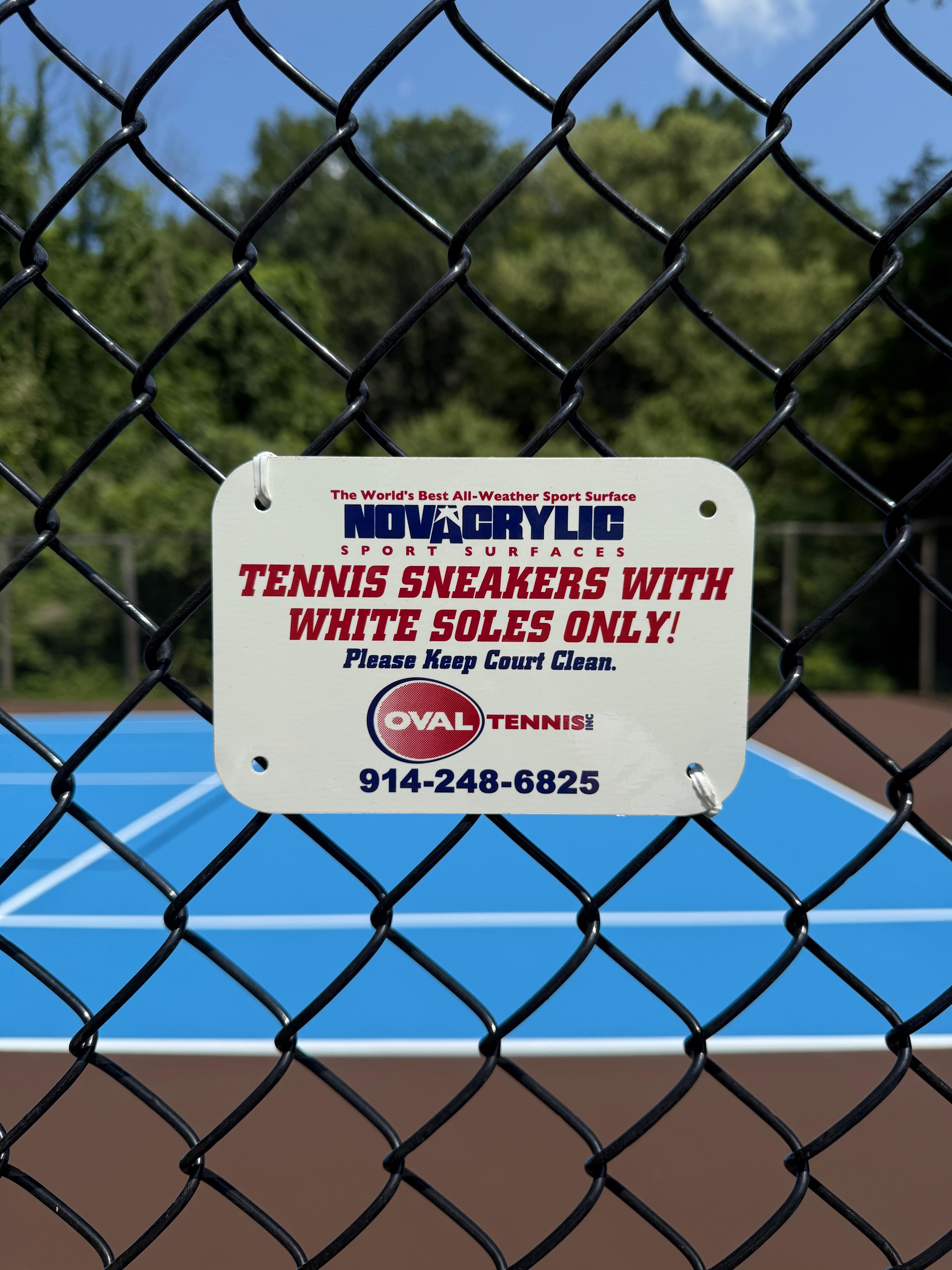 A white sign with red and blue text, 'NOVACRYLIC SPORT SURFACES TENNIS SNEAKERS WITH WHITE SOLES ONLY! Please Keep Court Clean!', is prominently displayed on a black chain-link fence, with a blue tennis court visible in the background.