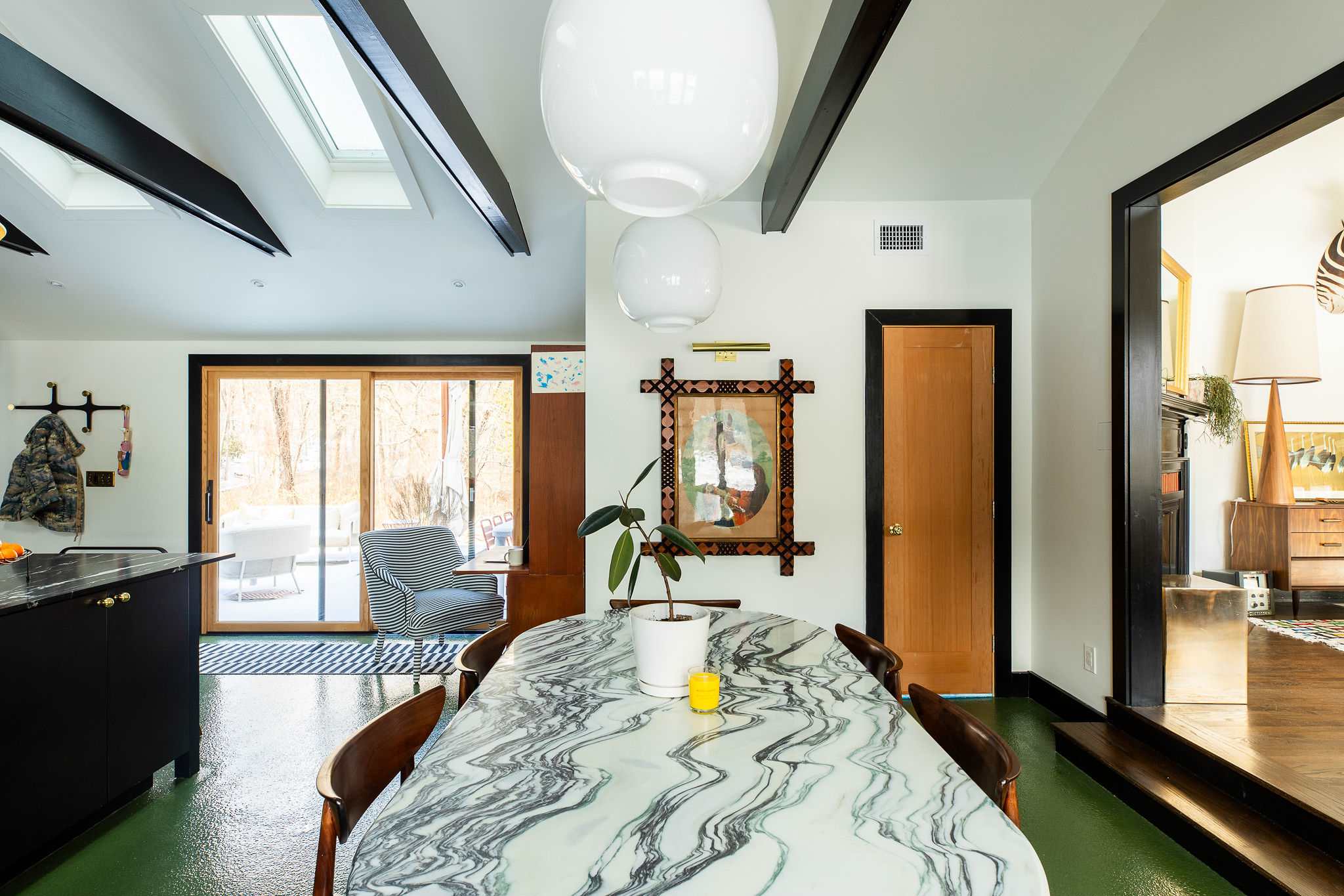A long, oval marble dining table with wooden chairs is centered under two large white pendant lights, with a view towards a sliding glass door leading to an outdoor area.