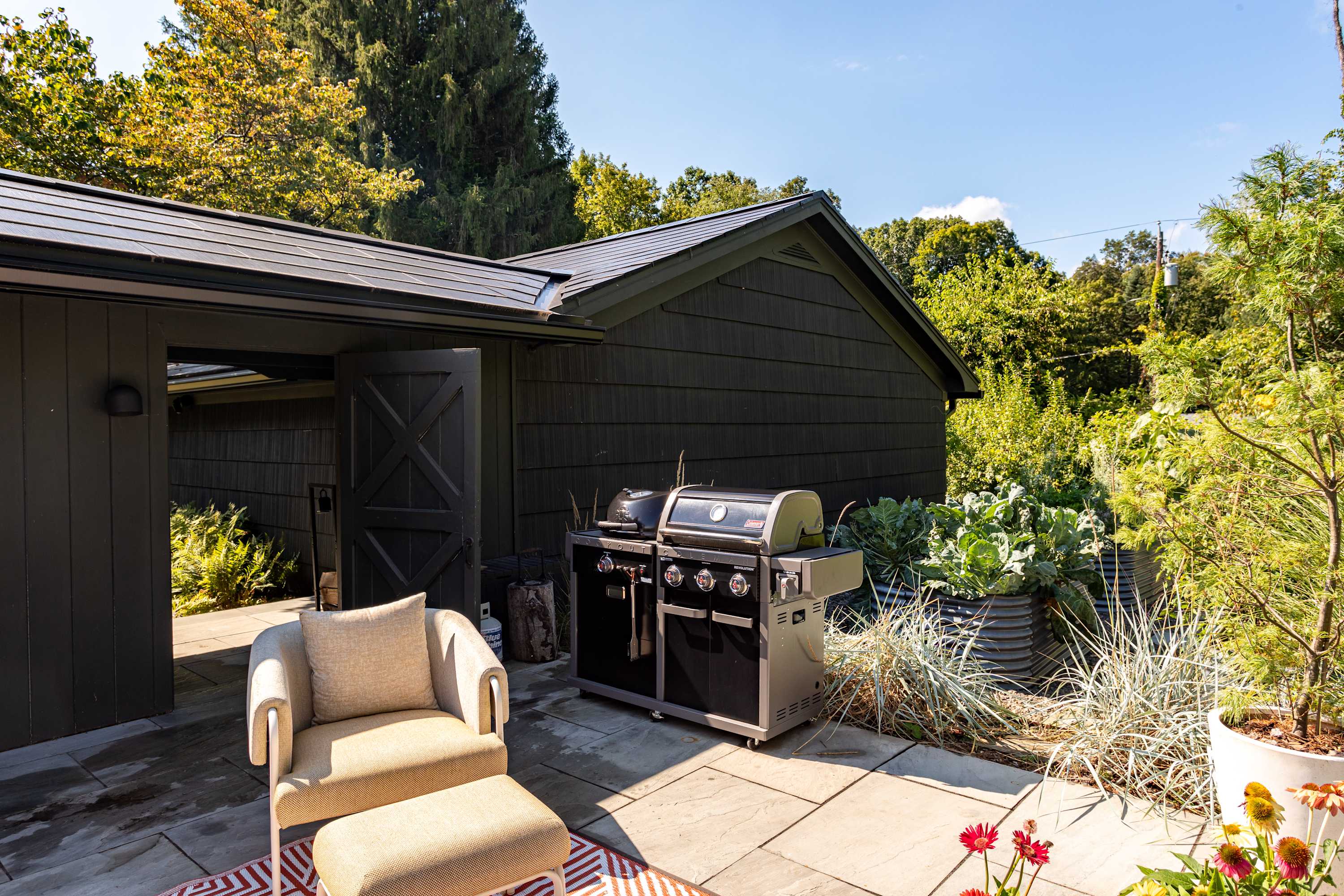 An outdoor patio area with a large black BBQ grill next to a dark-sided building, featuring a light beige armchair and ottoman on a stone patio, surrounded by various potted plants and lush greenery.
