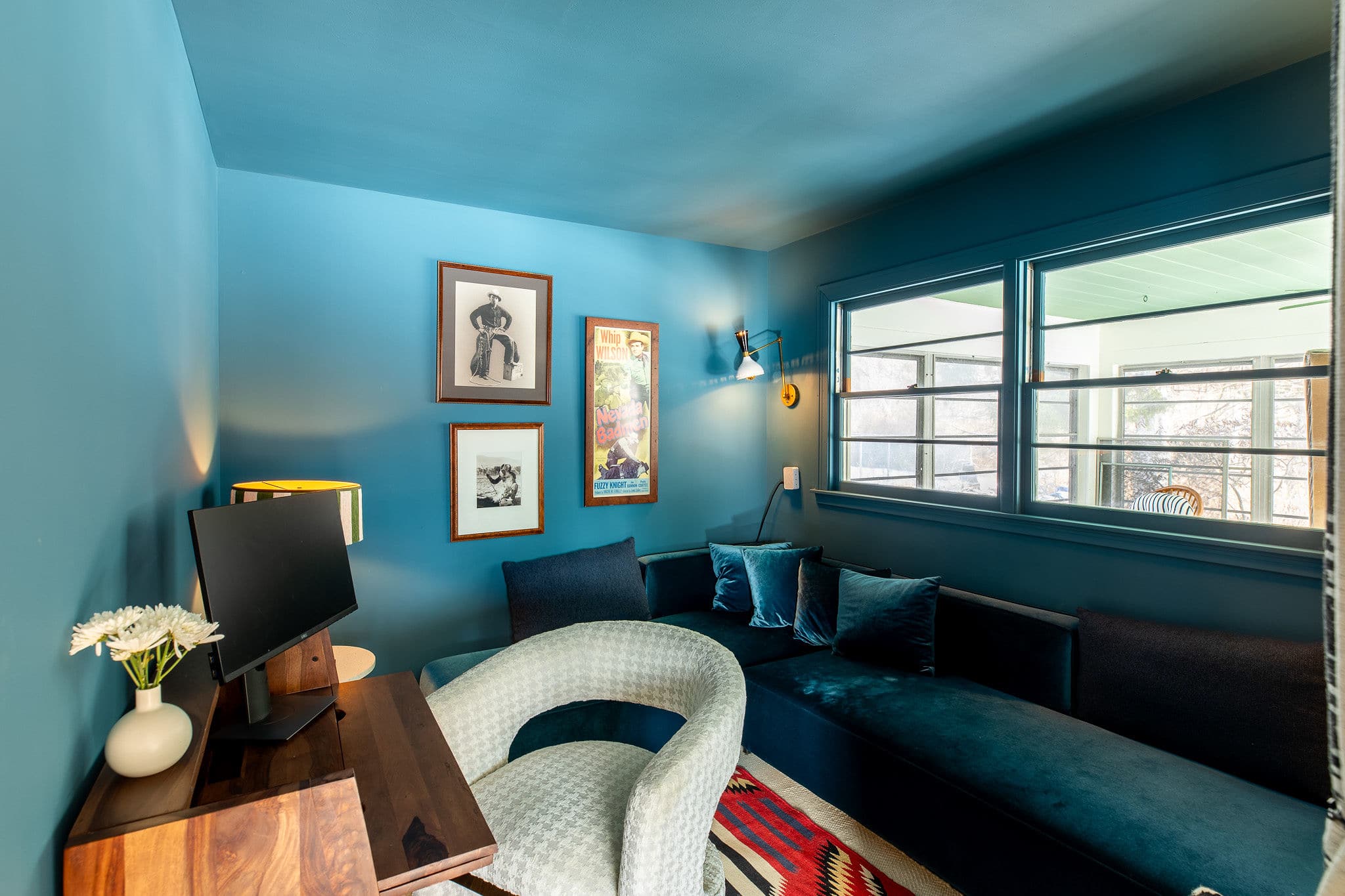 Cozy office space with deep teal walls and trim, furnished with a dark blue sectional sofa, a wooden desk with a monitor, and a white textured armchair. Artwork adorns the wall, and a window offers natural light.