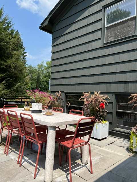 An outdoor dining area on a stone patio, featuring a large rectangular table with a speckled surface and several red metal chairs. The dark-sided house exterior and lush plants create a serene backdrop.