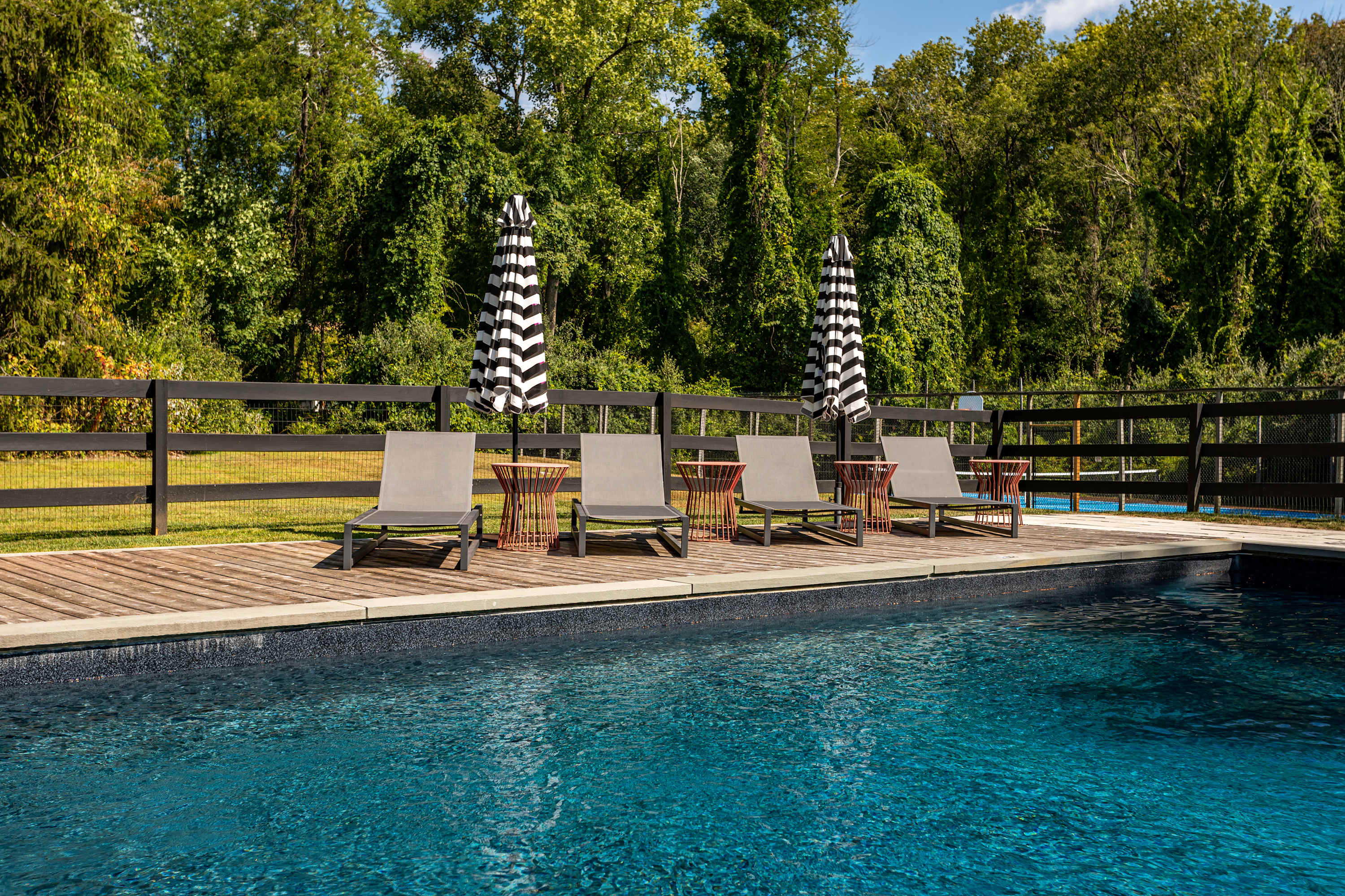 A serene dark blue swimming pool is bordered by a wooden deck with grey lounge chairs and striped umbrellas, offering a clear view of the surrounding green lawn, black fence, and dense forest beyond.
