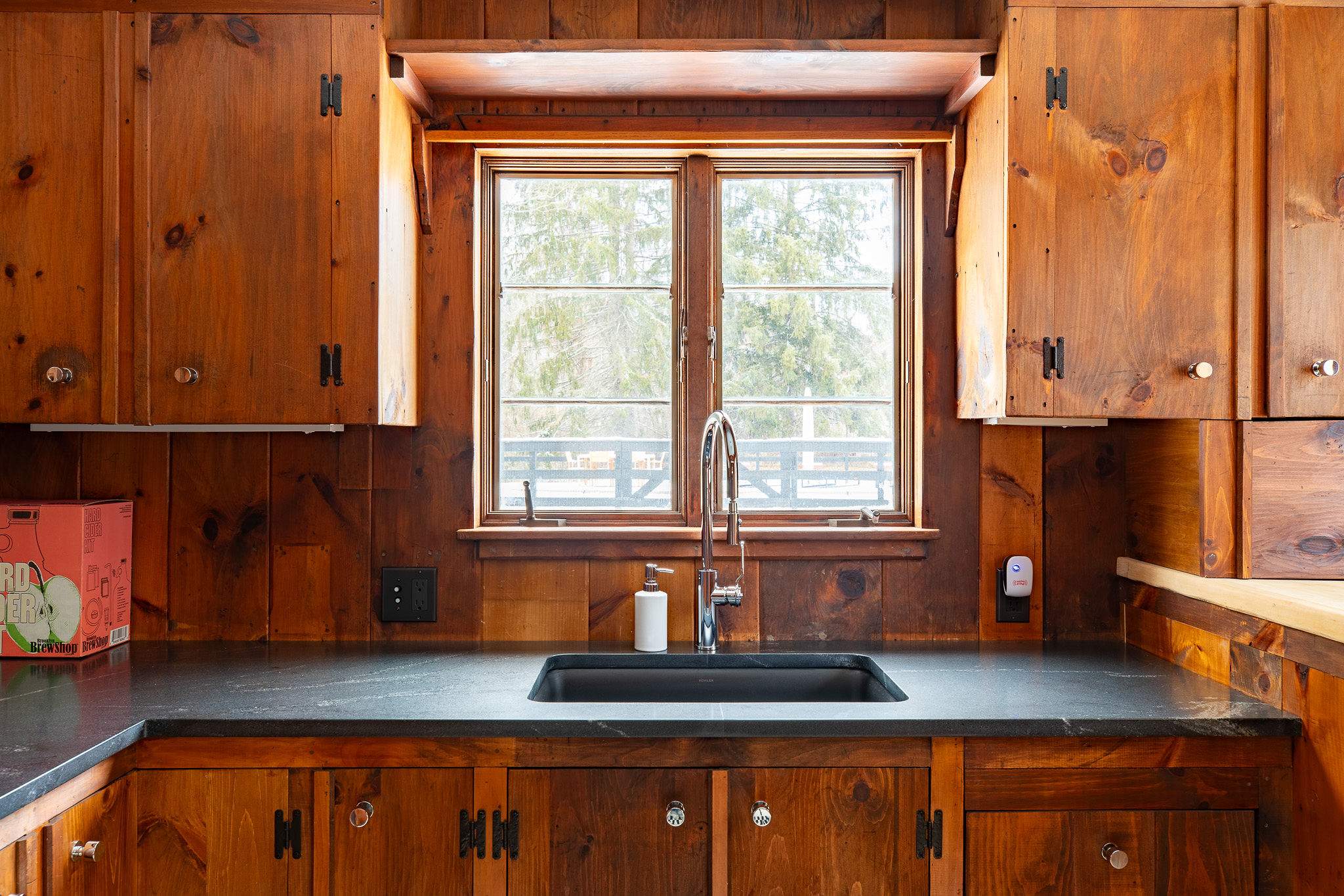 Kitchen area with rustic wood cabinetry and walls, featuring a dark countertop and a stainless steel sink beneath a large window offering views of the outdoors. A modern faucet completes the setup.