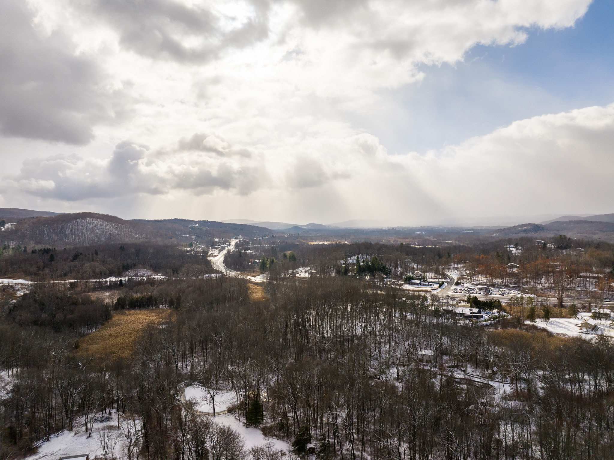 An aerial view of a vast winter landscape with snow-dusted trees and rolling hills under a dramatic cloudy sky with sun rays breaking through. A road winds through the valley, with distant buildings visible.