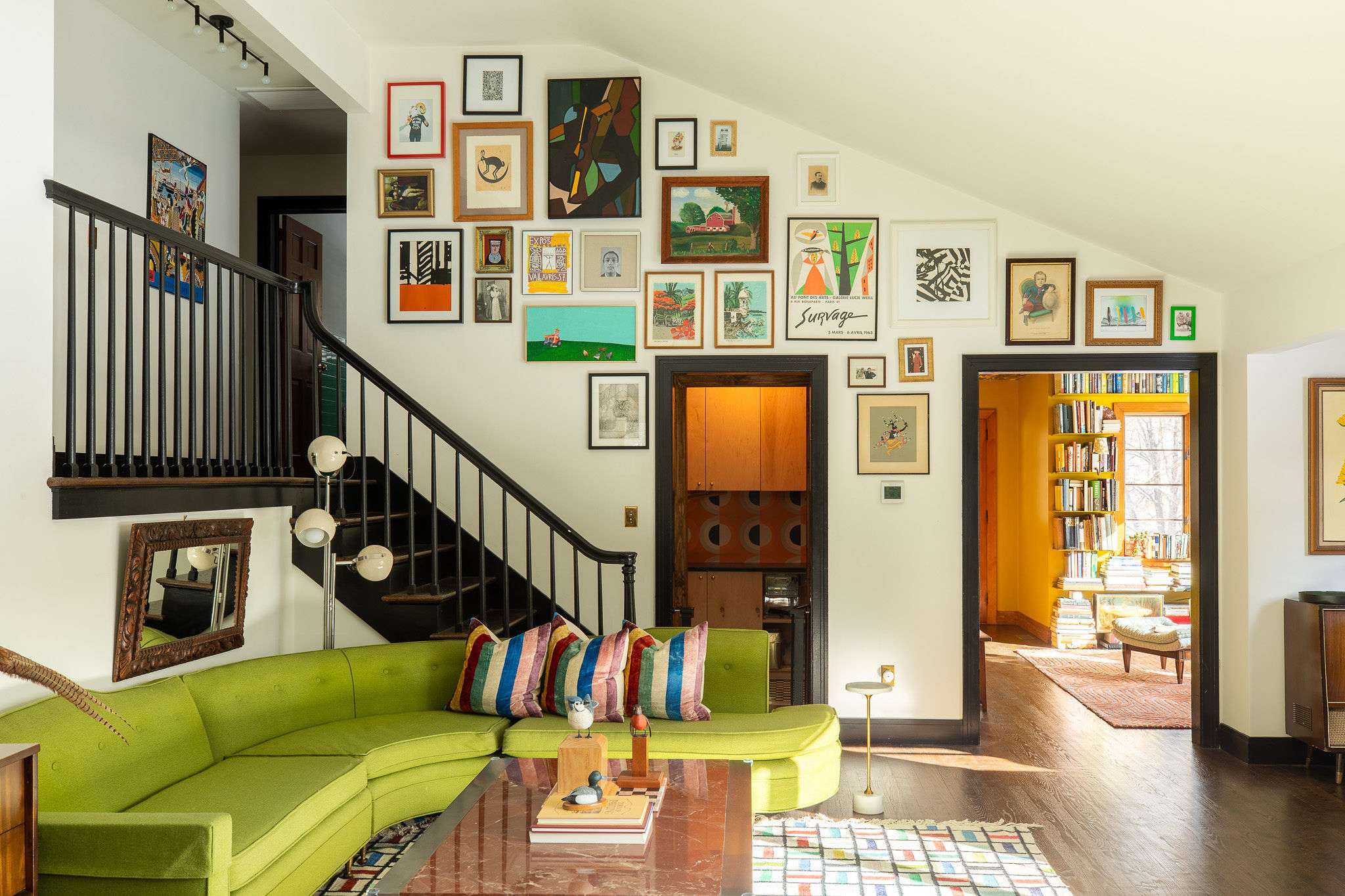 Vibrant living room featuring a lime green sectional sofa with colorful throw pillows, a dark wood coffee table, and a striking gallery wall filled with diverse artwork ascending alongside a black staircase.