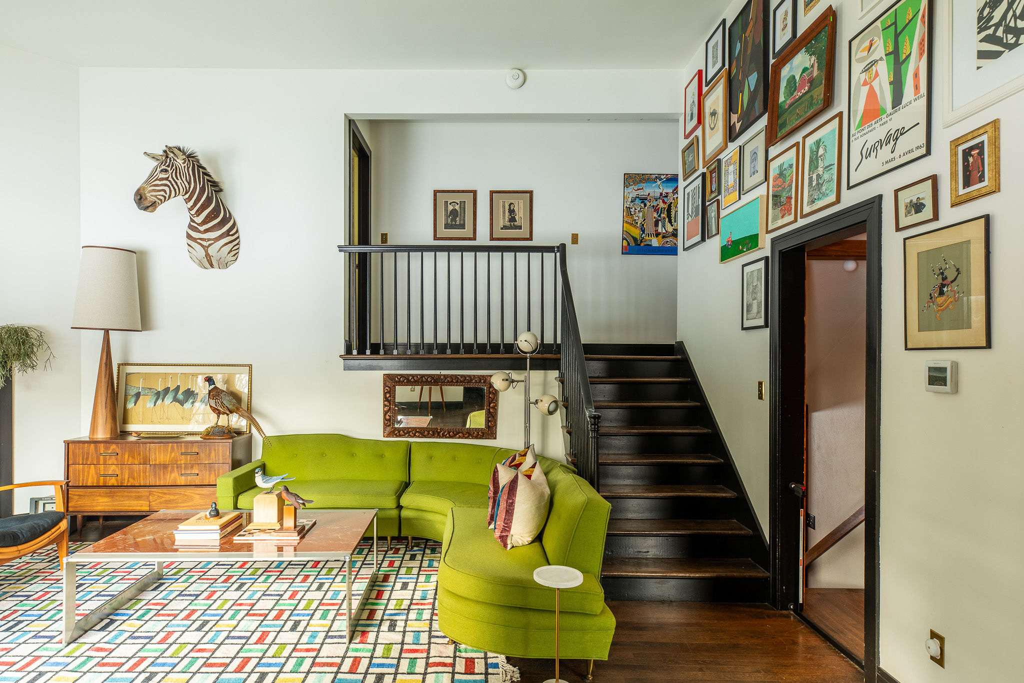 A vibrant living room features a large, curved lime green sectional sofa on a colorful geometric rug. A gallery wall of diverse artwork extends up the staircase, which has dark wood steps and a black railing. A zebra head mount adds a quirky touch.