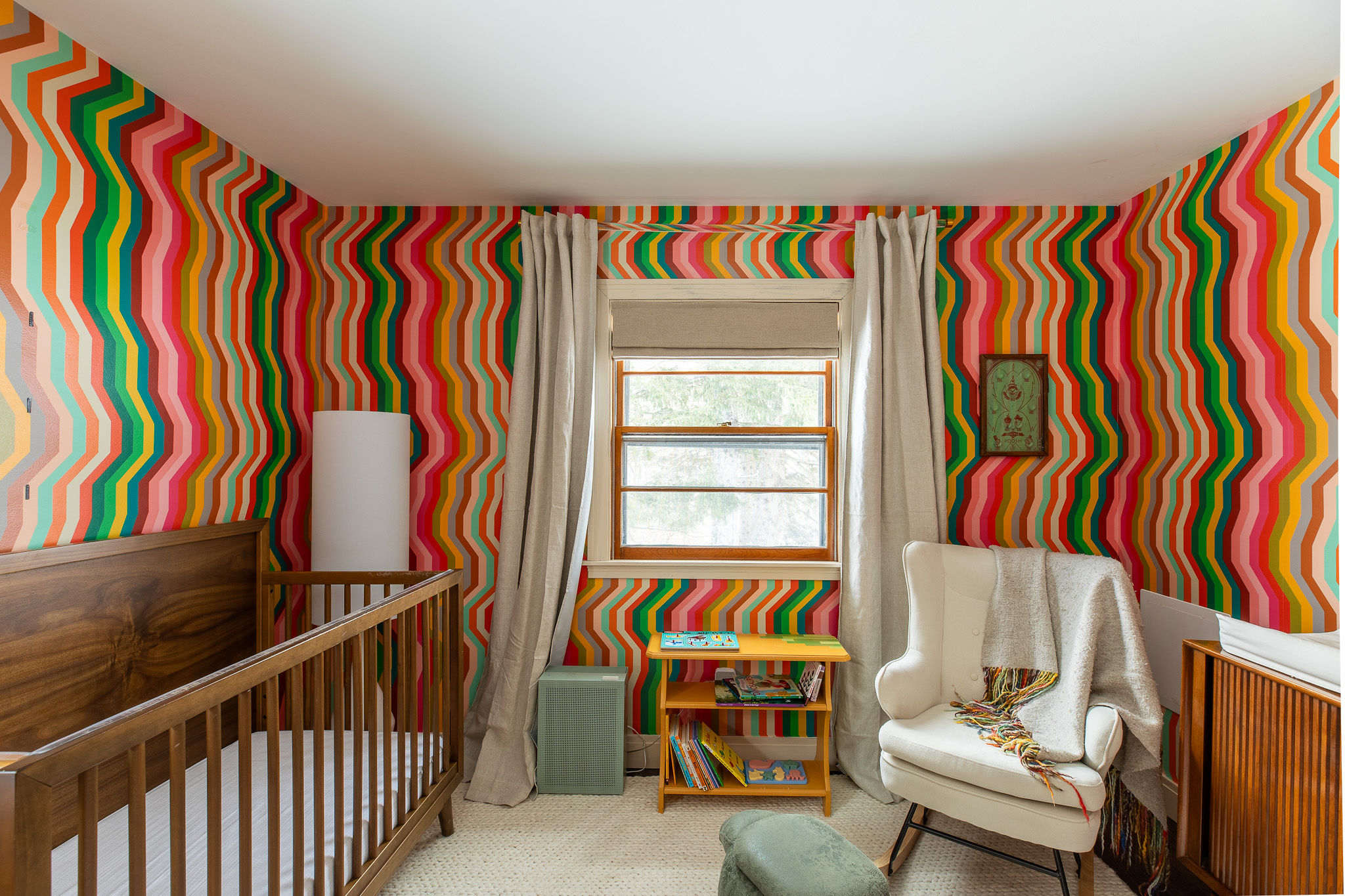 Vibrant nursery with a bold, colorful striped wallpaper in shades of red, orange, green, and pink, featuring a wooden crib, a white armchair, and a small wooden bookshelf. A window with light curtains provides natural light.