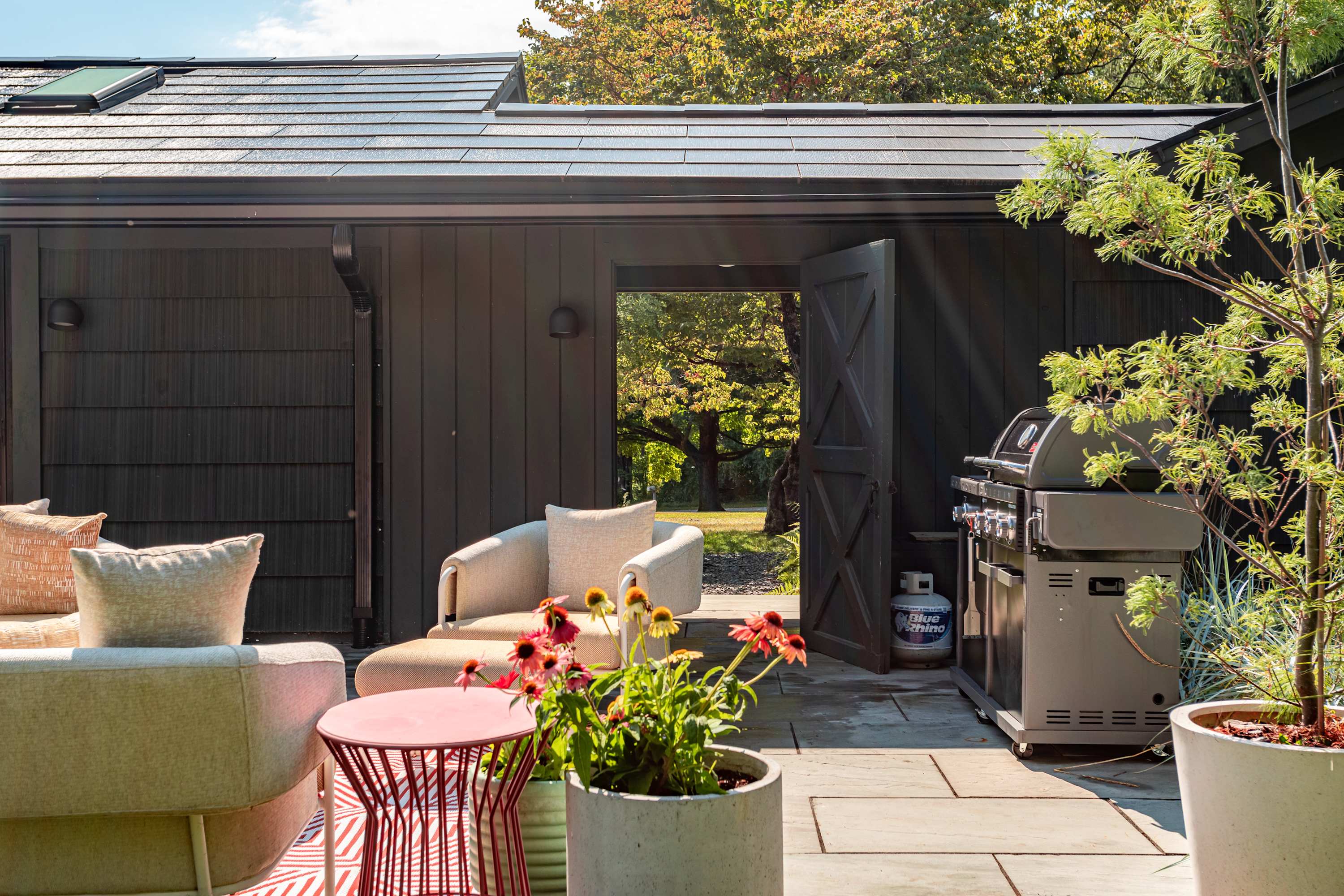 A cozy outdoor patio features comfortable upholstered seating, a pink metal side table, potted plants with flowers, and a large BBQ grill, all situated on stone pavers next to a dark-sided building with an open doorway.