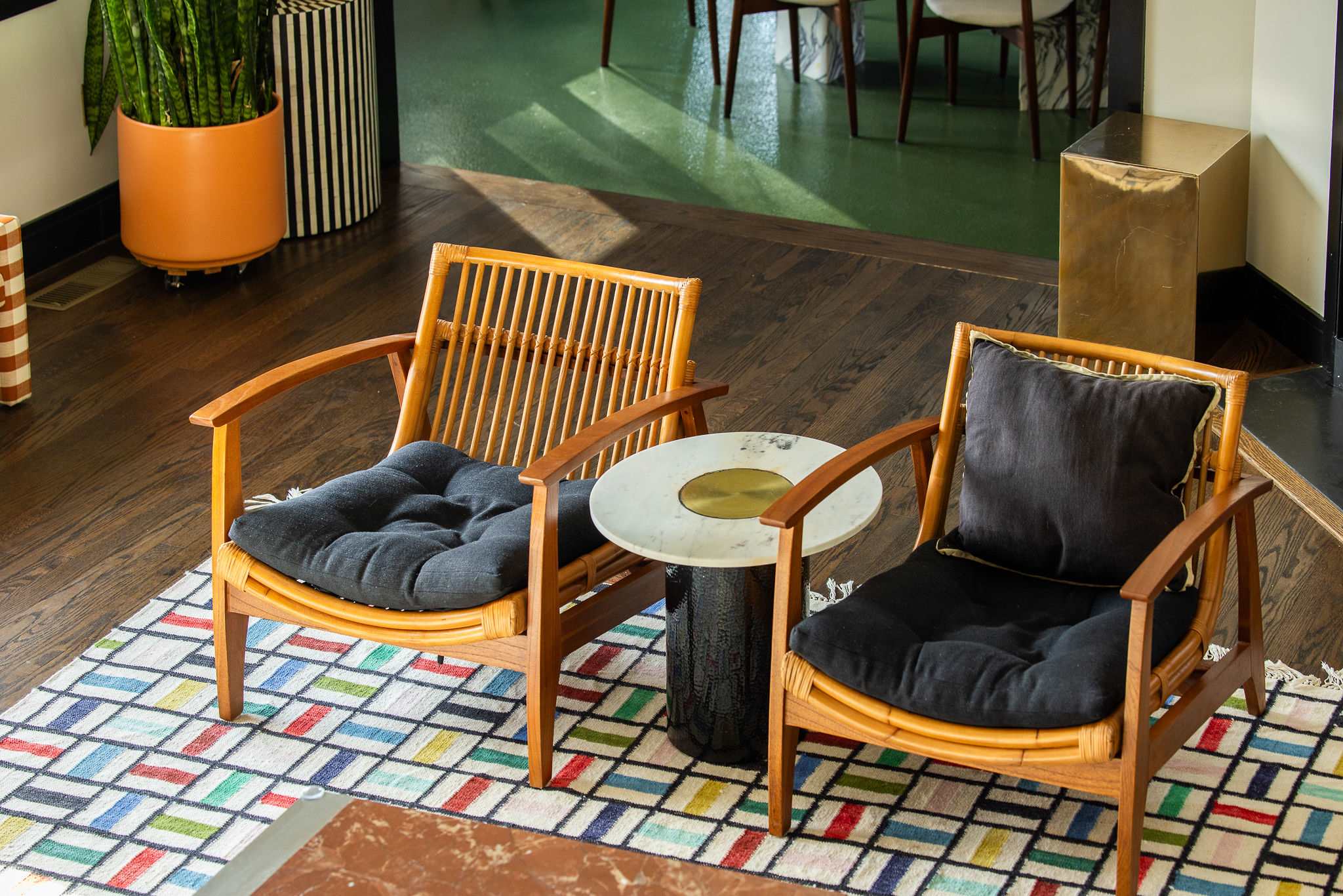 Two rattan armchairs with dark cushions flank a small round marble-topped side table, all resting on a colorful geometric rug with a dark wood floor visible around it.